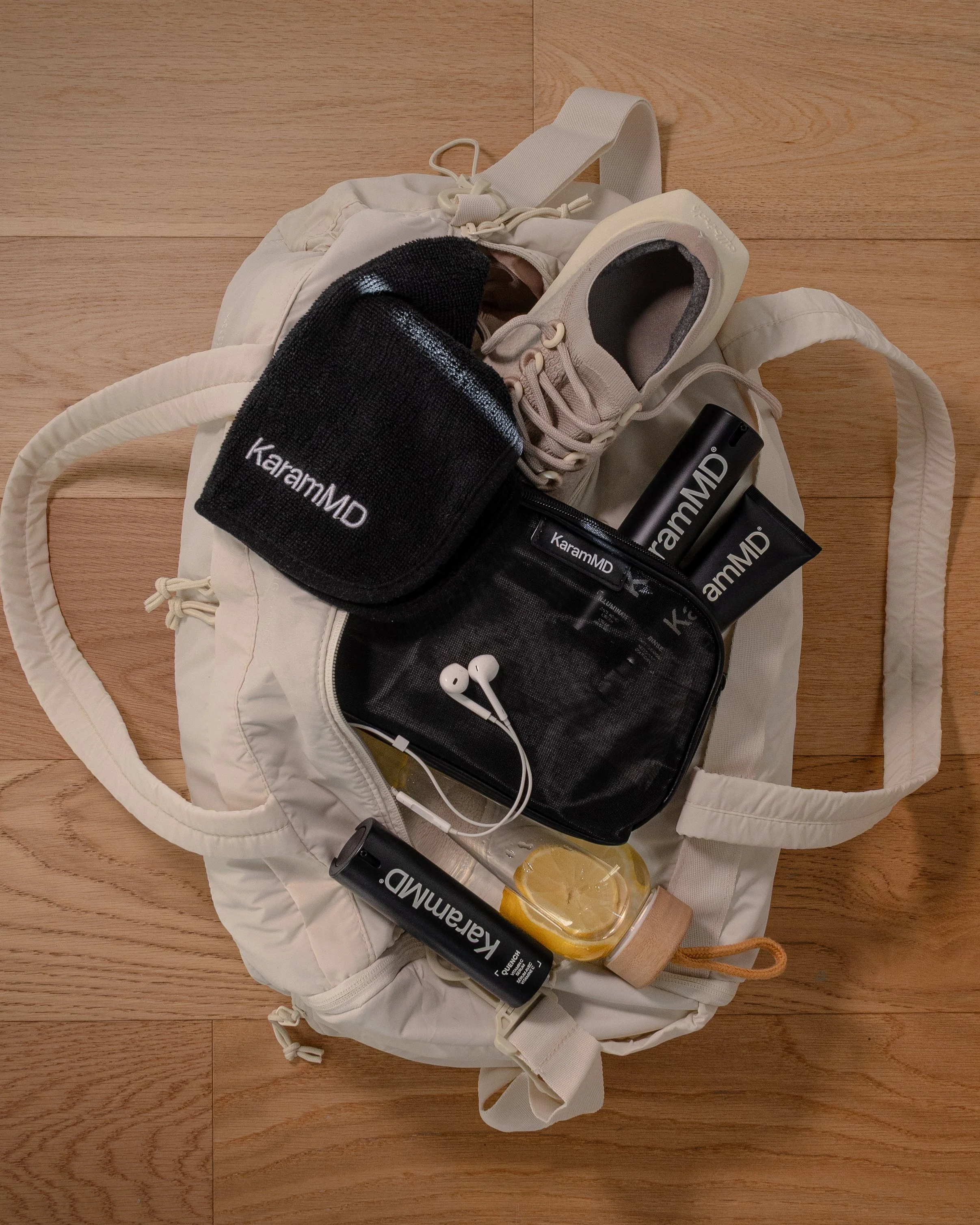 A white sports bag on a wooden floor containing Sleep Mask, sneakers, KaramMD skincare products, Earphones, and Lemon water.