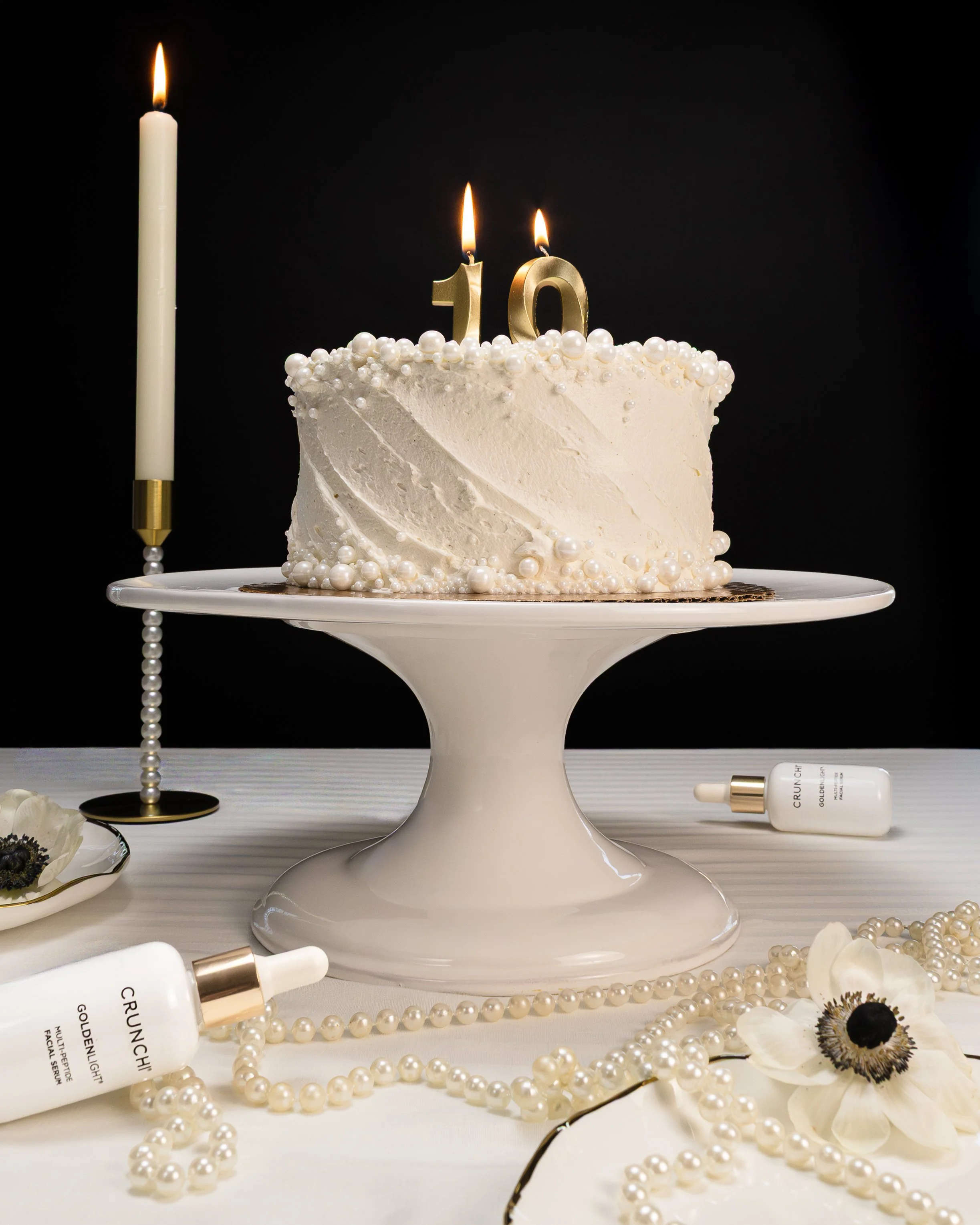 A white birthday cake with pearl decorations and gold number candles '10' on top, on a white cake stand. There are lit candles, pearl necklaces, skincare bottles, and white flowers around the cake on a white table with a black background.