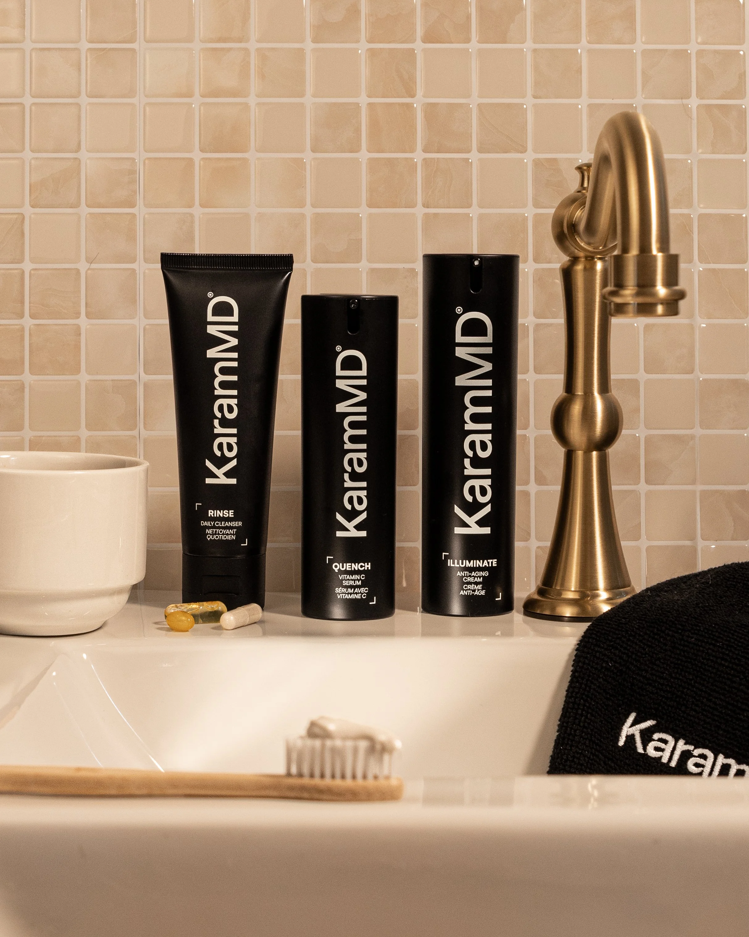 KarameMD skincare products on a bathroom sink counter, including rinse, quench, and illuminate, alongside a white bowl, a black towel with the brand name, a toothbrush with toothpaste, a toothbrush capsule, and a gold-colored faucet, with small beige