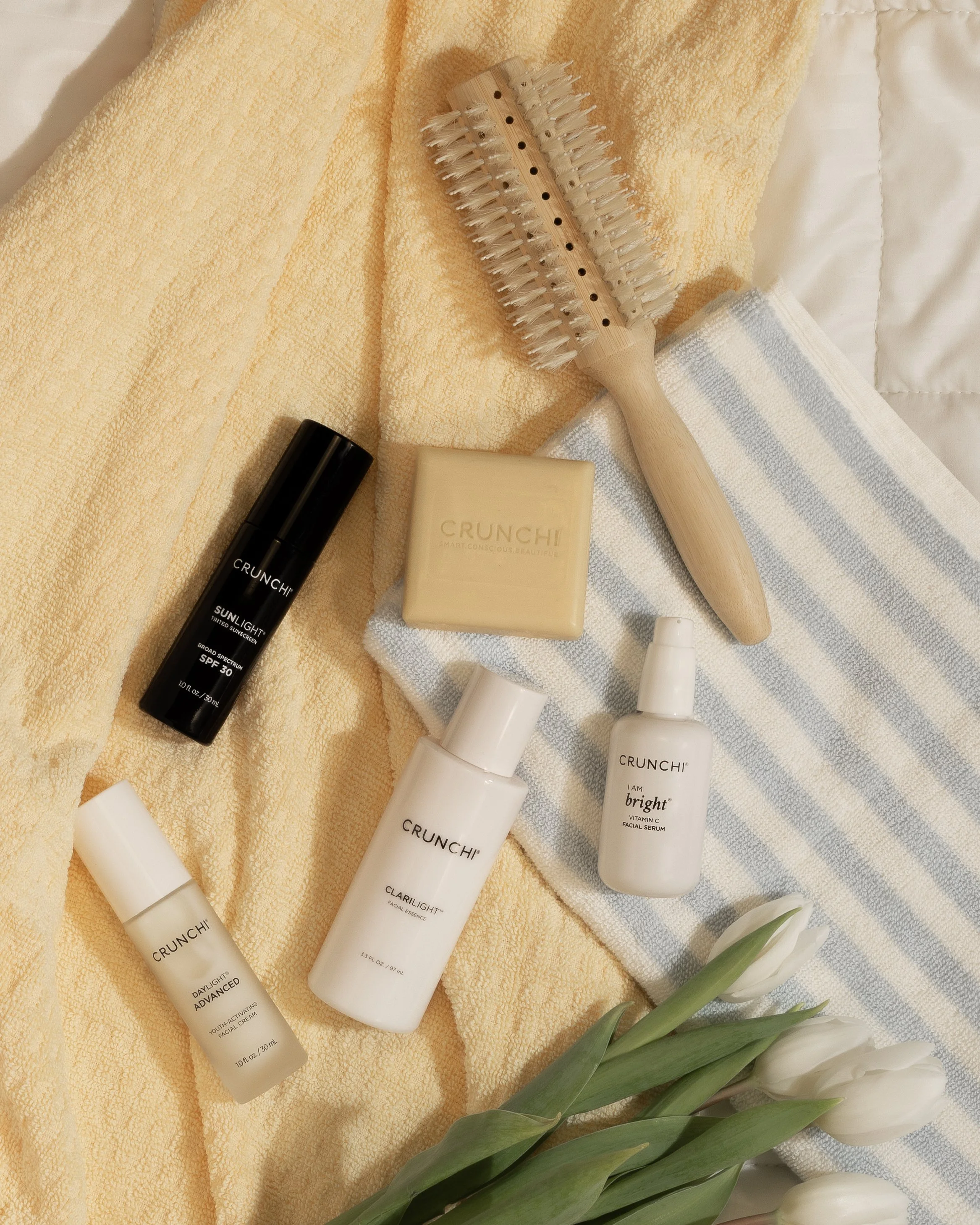 Skincare and beauty products arranged on a soft yellow towel and striped cloth, including a black SPF sunscreen, a bar soap, facial serum, face cream, and a facial essence, with a wooden massage brush and white flowers nearby.