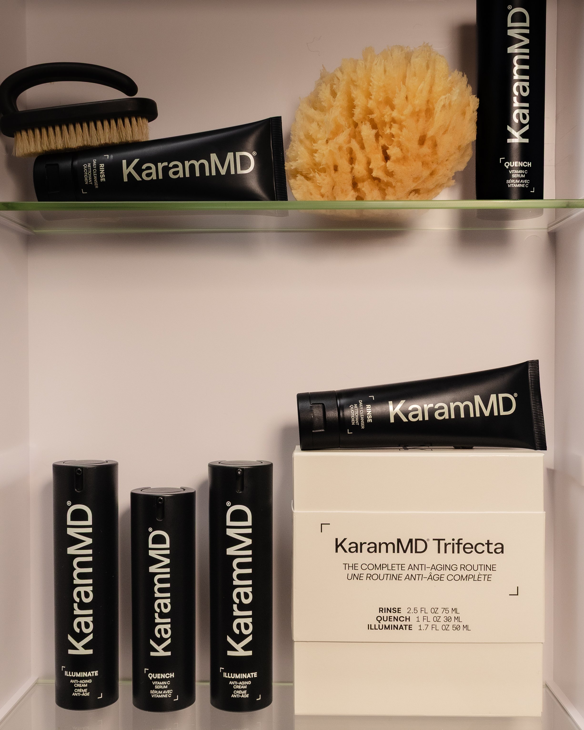 Skincare products from KaramMD, including tubes labeled 'KaramMD,' a box labeled 'KaramMD Trifecta,' and black containers with product names such as 'Illuminate' and 'Quench,' along with a sponge and a brush.