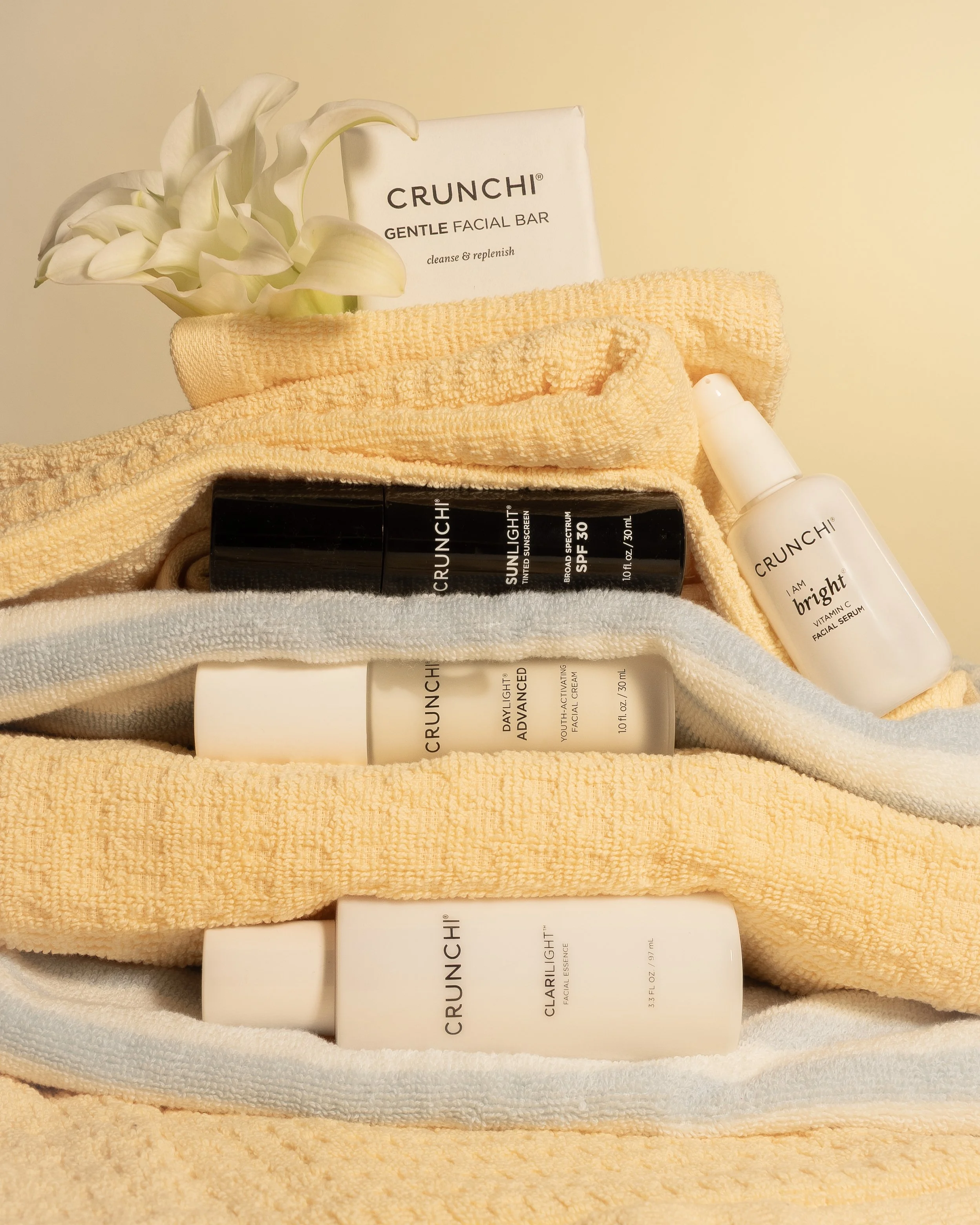 A collection of skincare products and towels, including a gentle facial bar, facial serums, broad spectrum SPF 30 sunscreen, and a white flower arrangement.