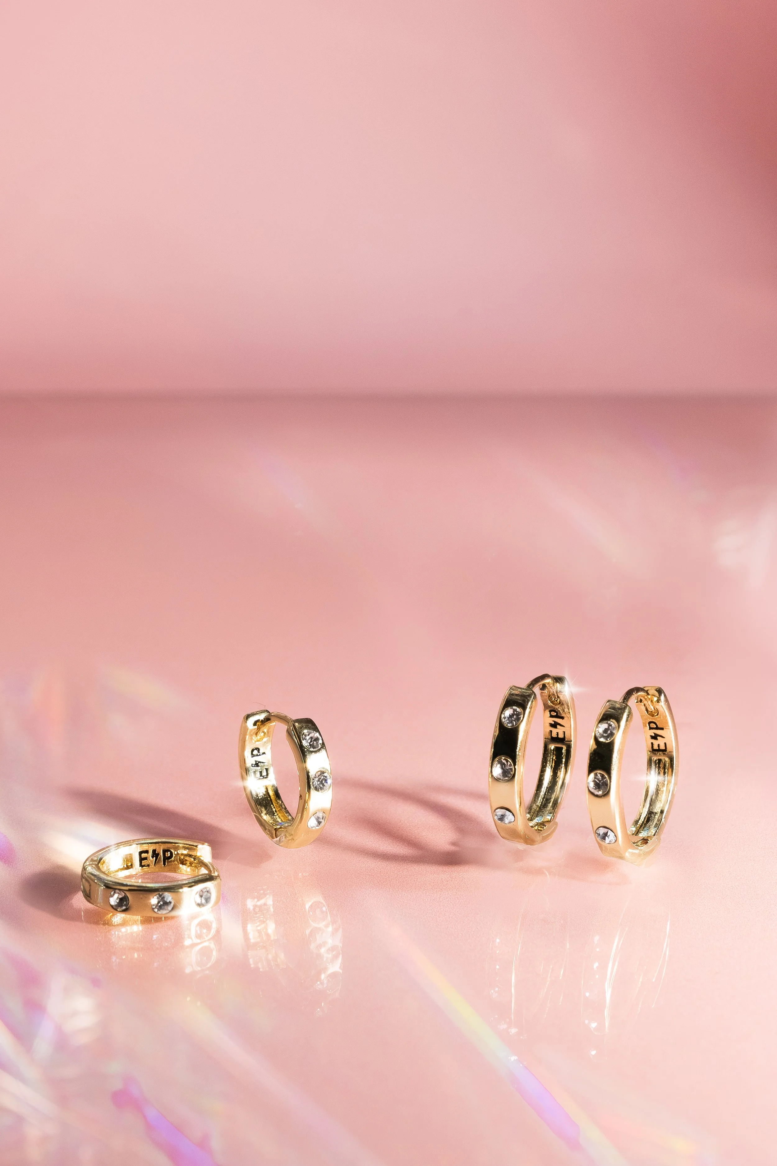 Set of five gold hoop earrings with embedded clear stones, displayed on a pink reflective surface with a pink background.