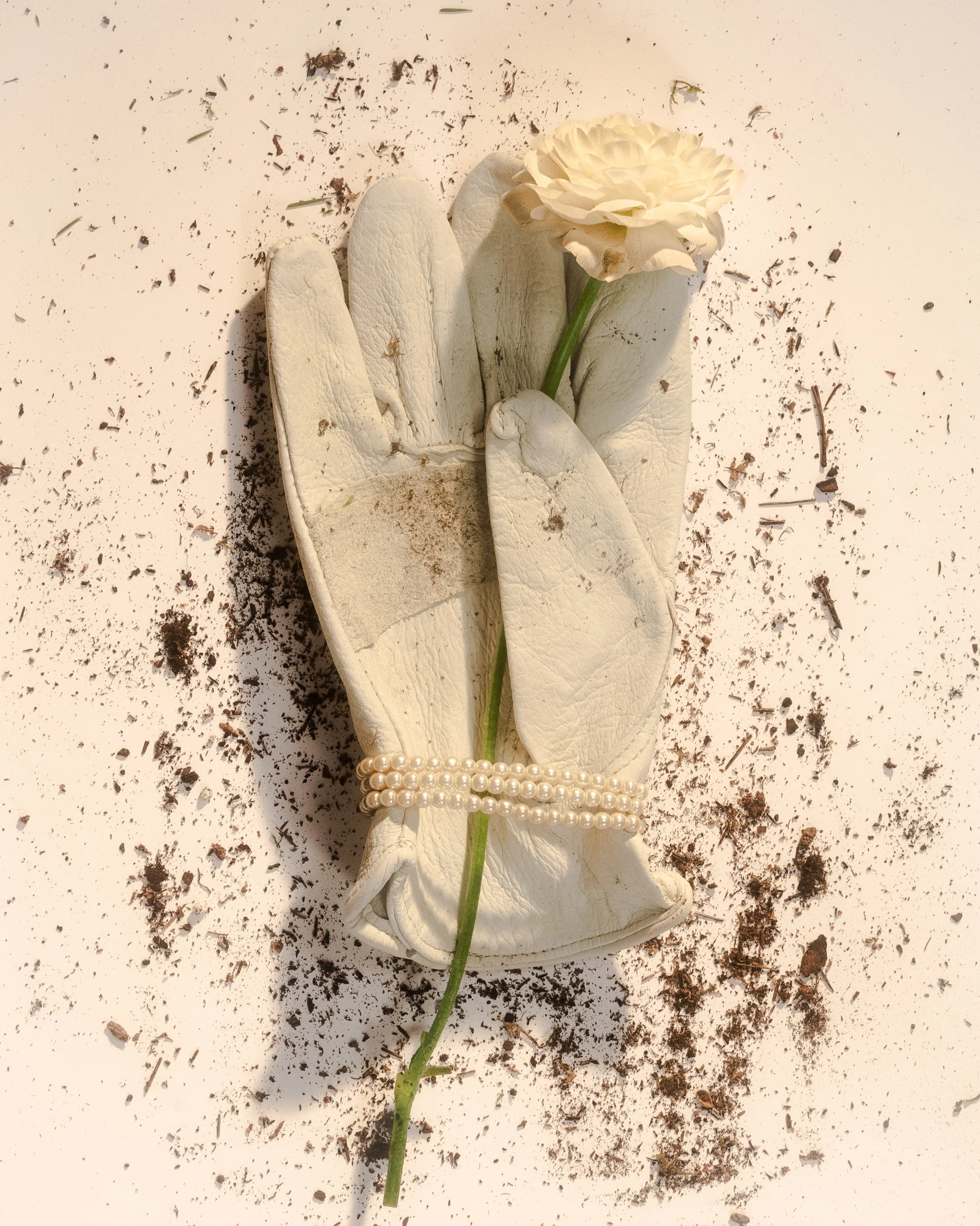 A gardening glove with a pearl bracelet on it holding a cream-colored flower, with soil scattered around on a white surface.