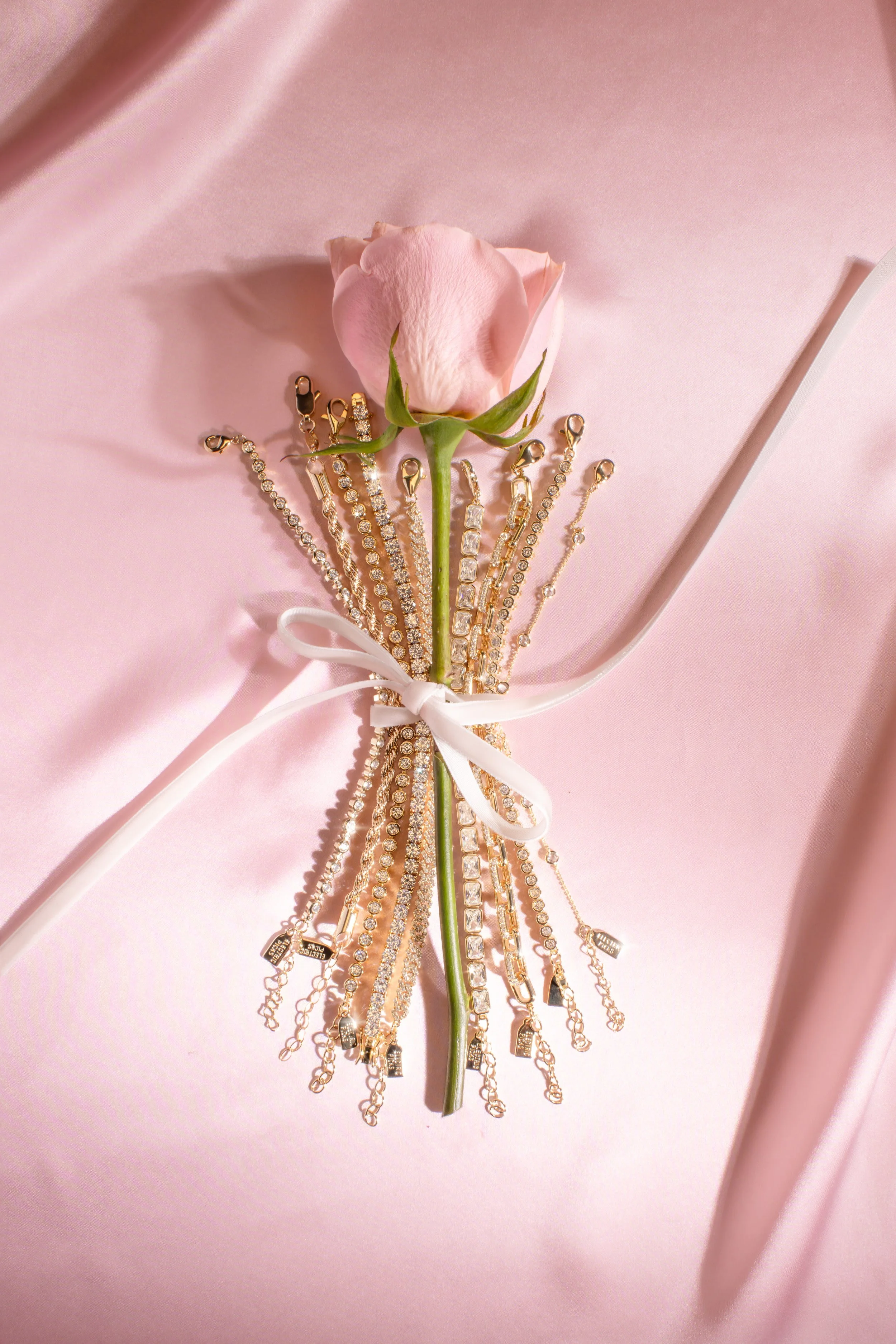 A pink rose with a white ribbon surrounded by multiple gold and diamond bracelets on a soft pink background.
