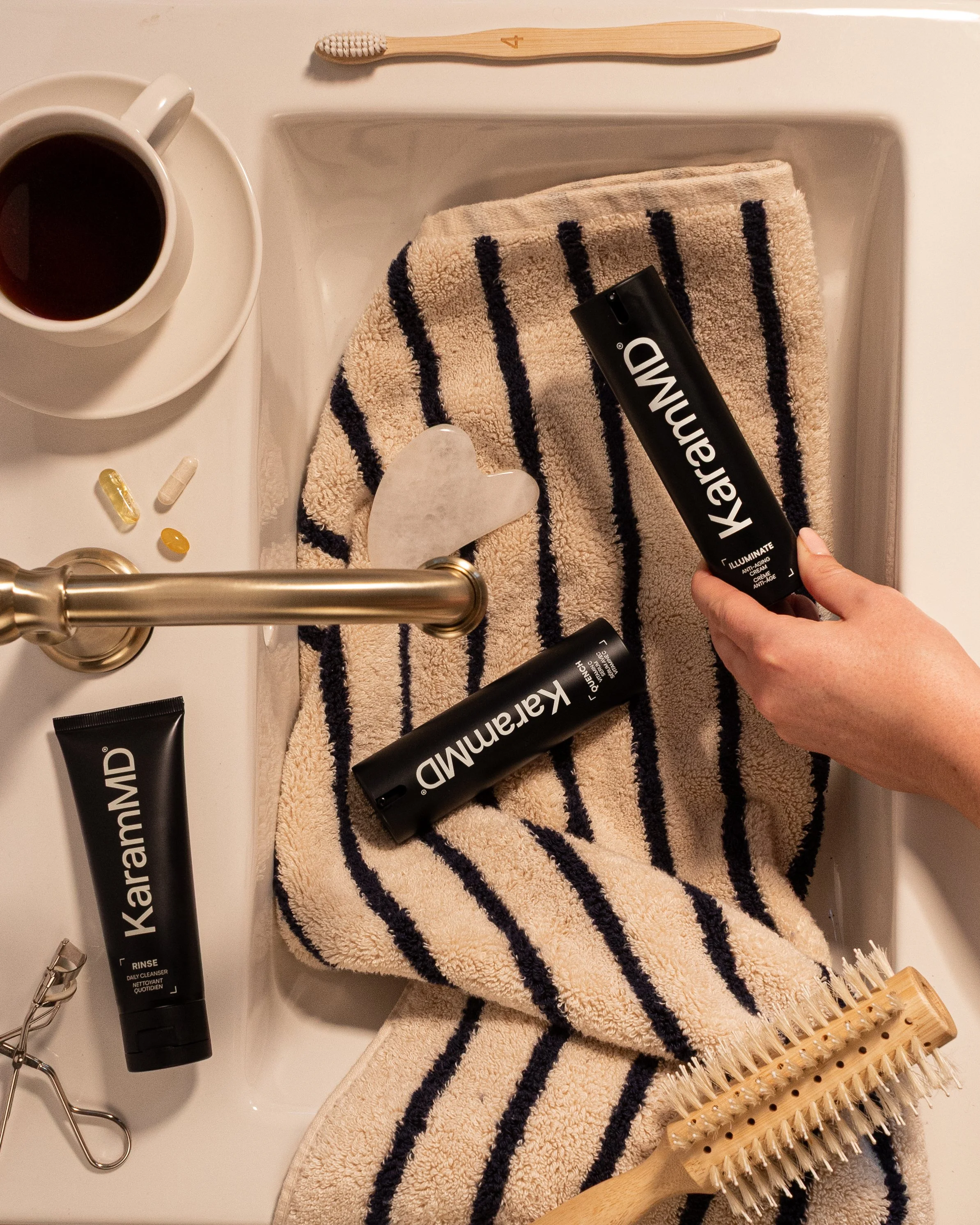 A sink filled with a striped towel, a wooden hairbrush, and two black tubes of Kaaramed skincare products. A hand is holding one of the tubes. Nearby, there is a coffee cup, some pills, a wooden toothbrush on top of the towel, and a pair of eyelash c