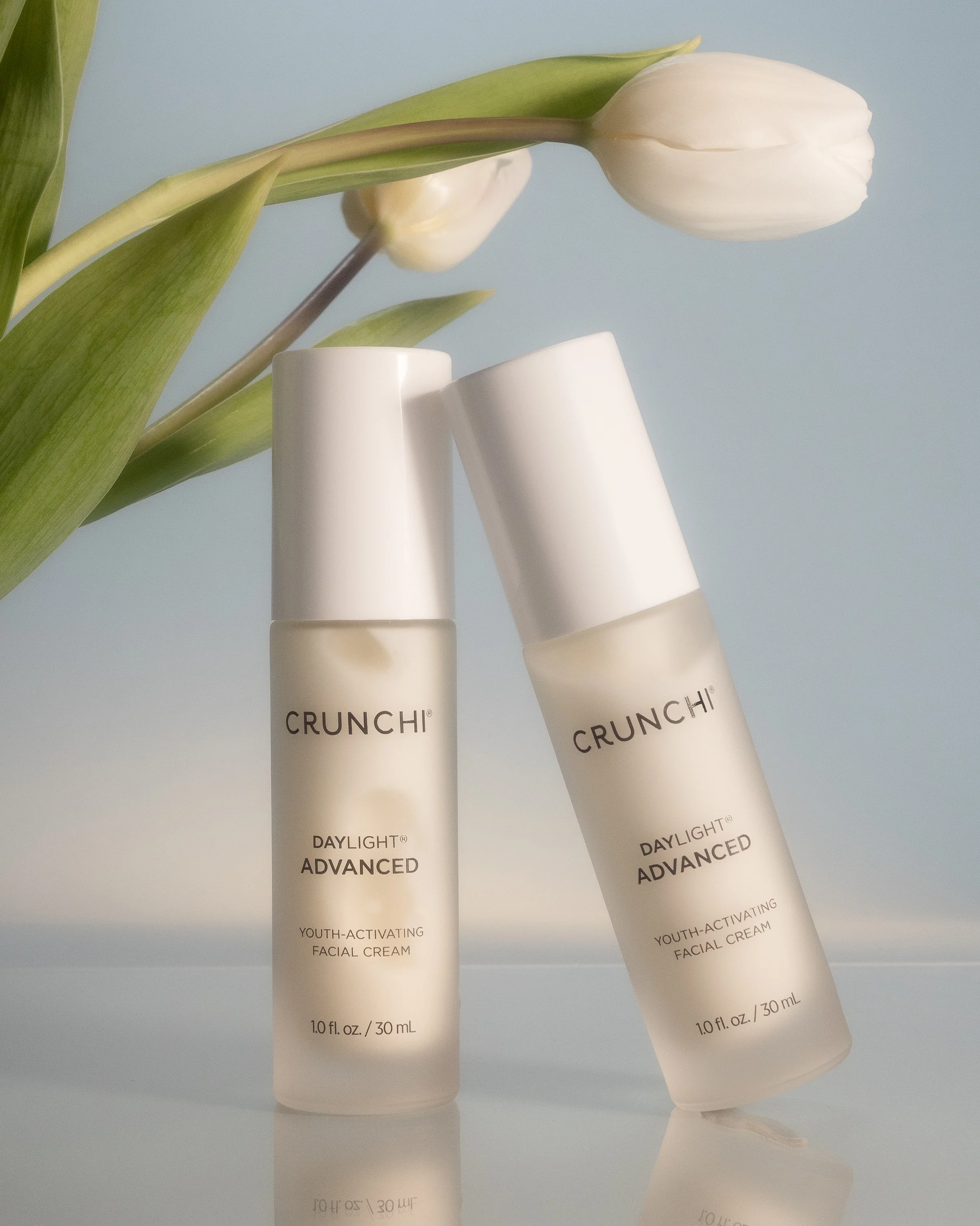 Two bottles of CRUNCHI DAYLIGHT ADVANCED youth-activating facial cream with white caps, set against a light blue background with a white tulip flower nearby.