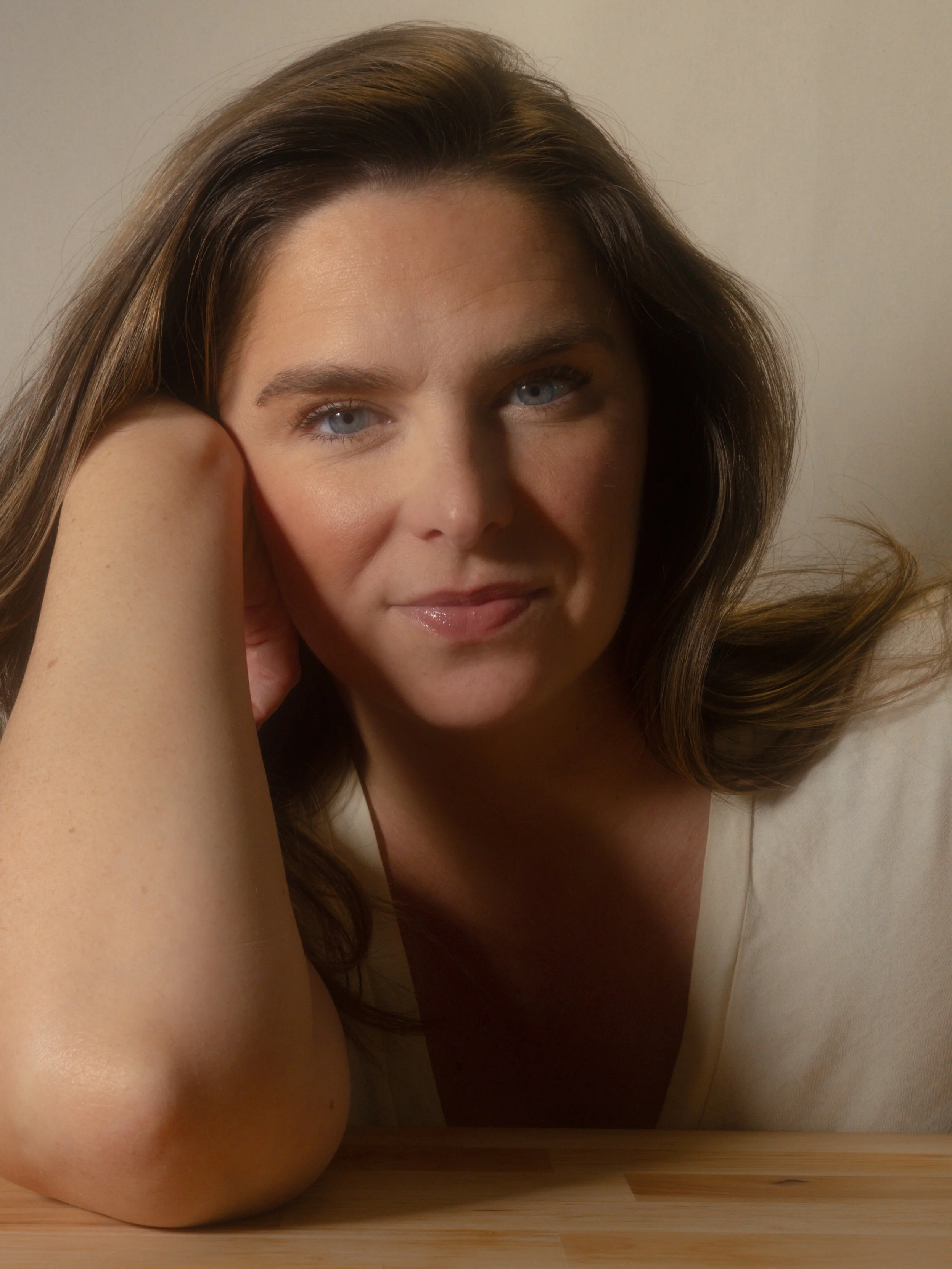 Close-up of a woman with long brown hair and blue eyes, resting her head on her arm, lying on a wooden floor near a wall.