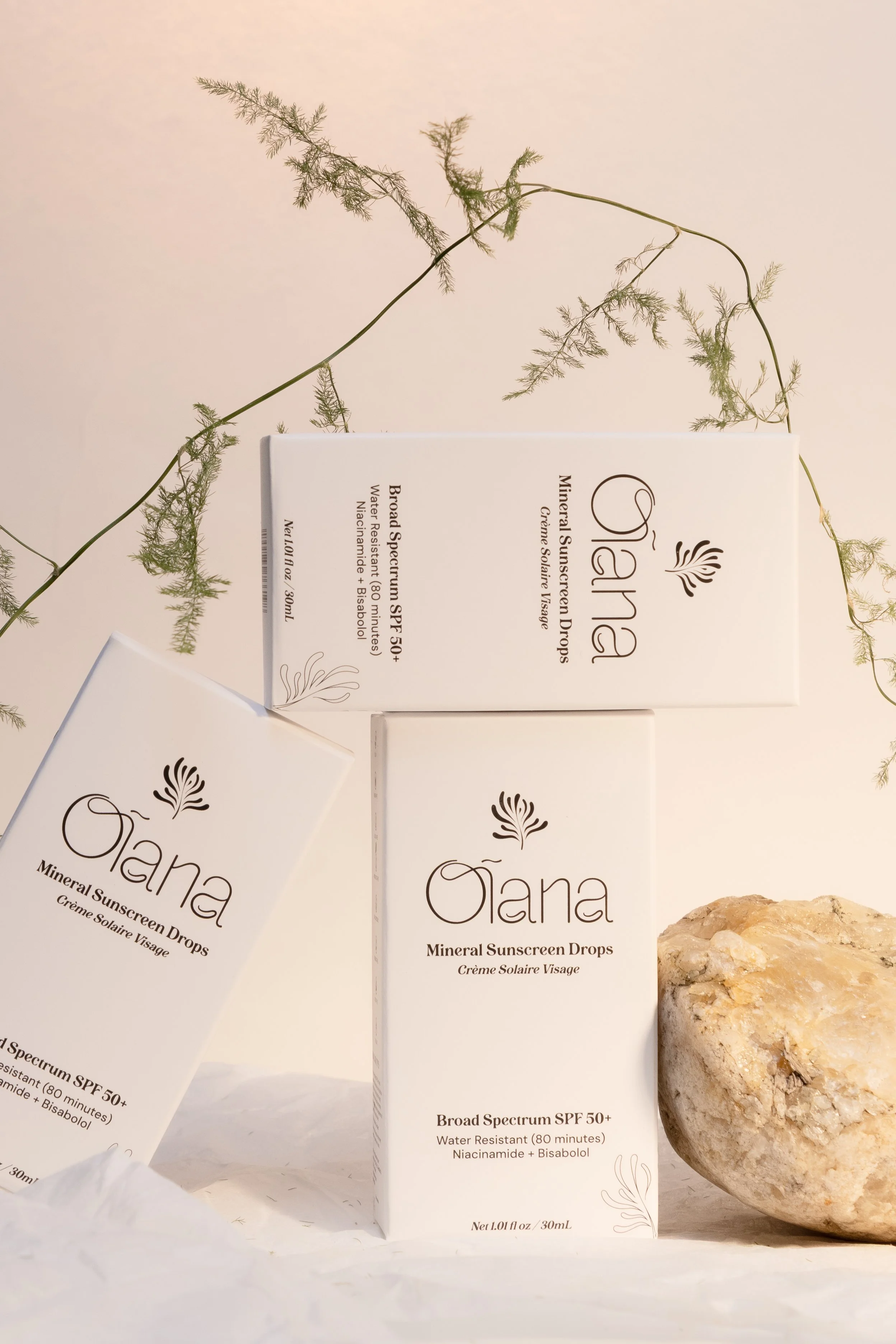 Three boxes of Olaf skincare products, two of which are standing while the other is leaning against a rock, with a delicate green plant in the background.