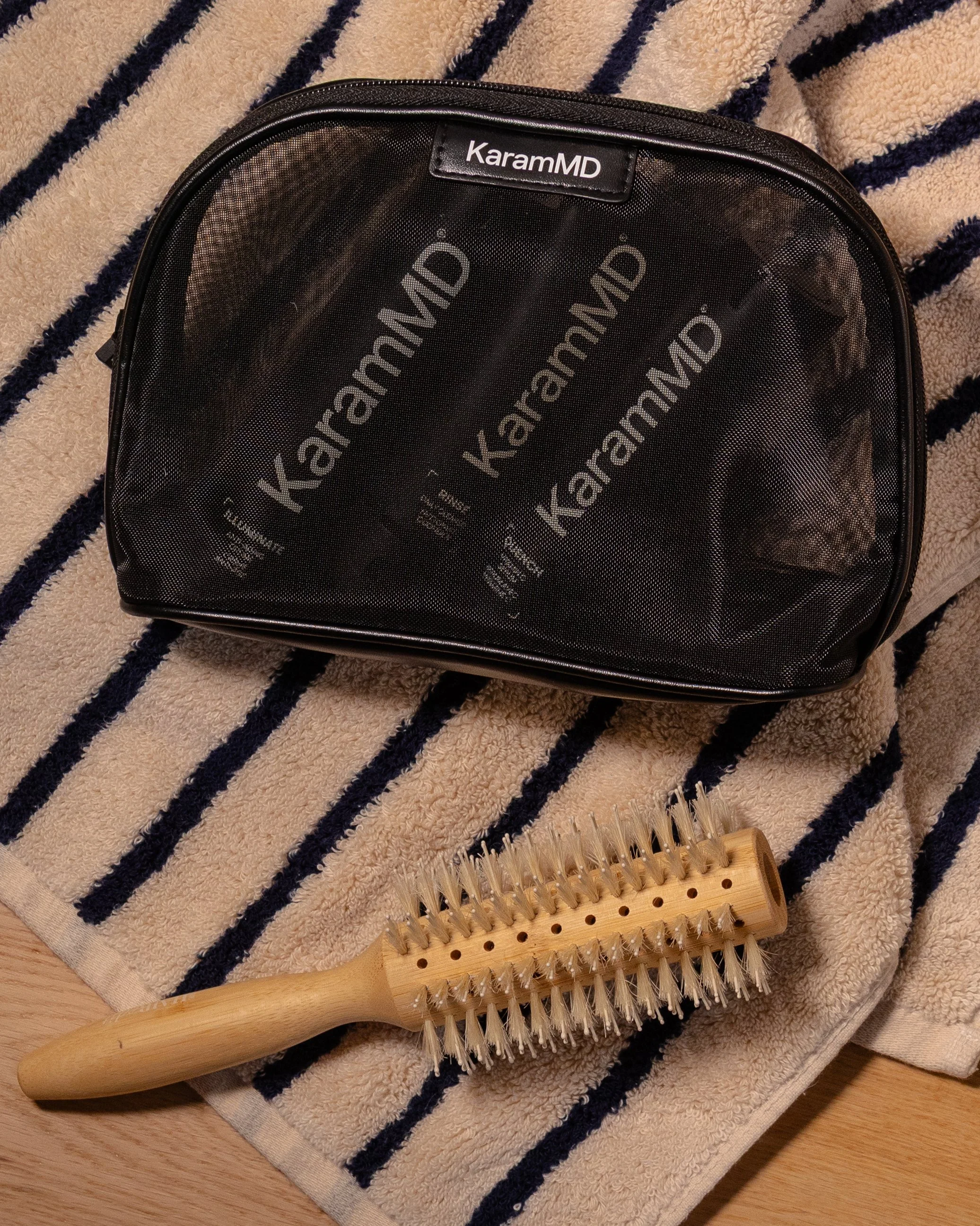 A black toiletry bag labeled 'KaramMD' with a clear plastic front, resting on a striped towel. A wooden massage roller with multiple small spiked rollers is also on the towel.