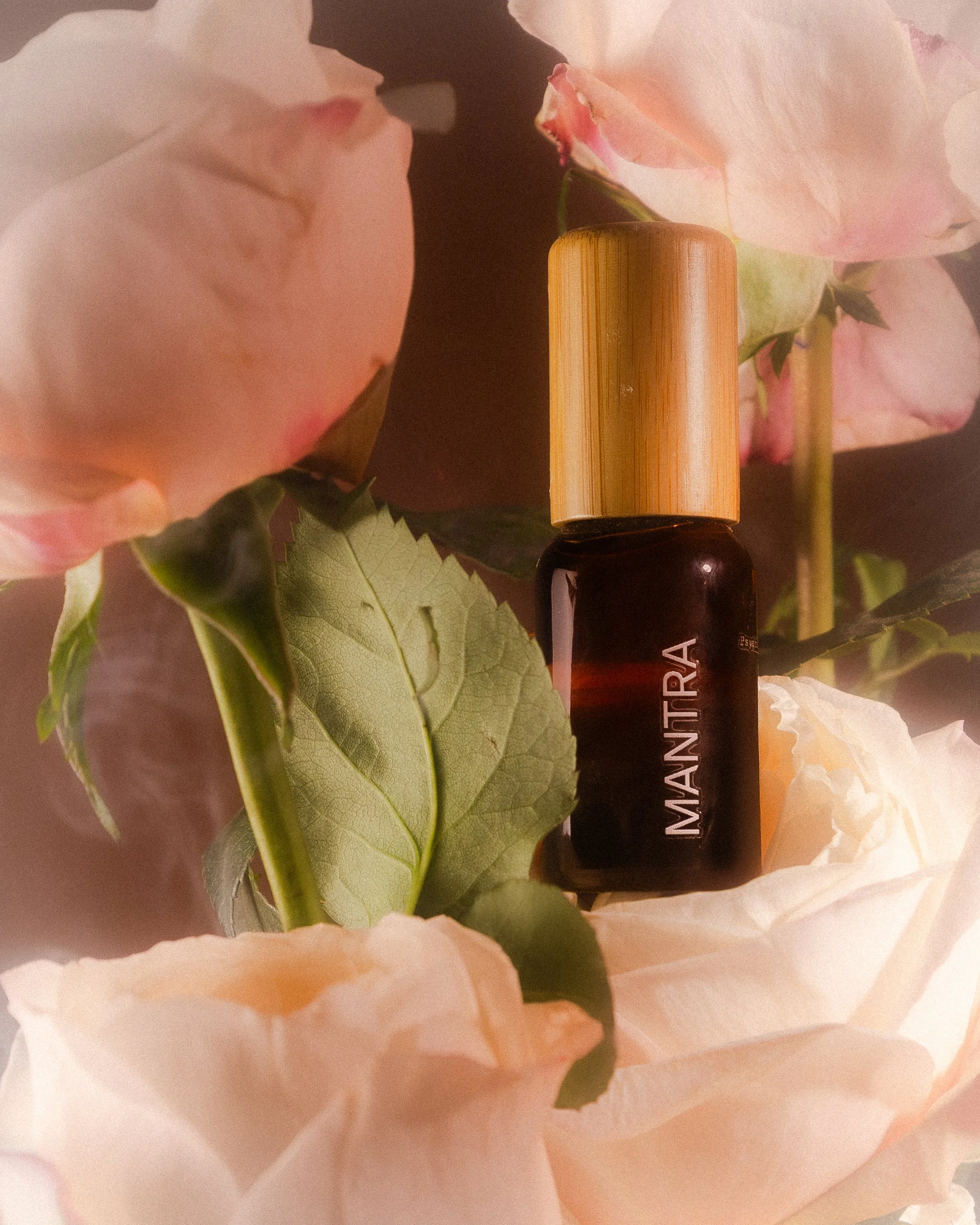 A small brown essential oil bottle with a golden cap labeled 'MANTIRRA', surrounded by pink and white roses and green leaves.