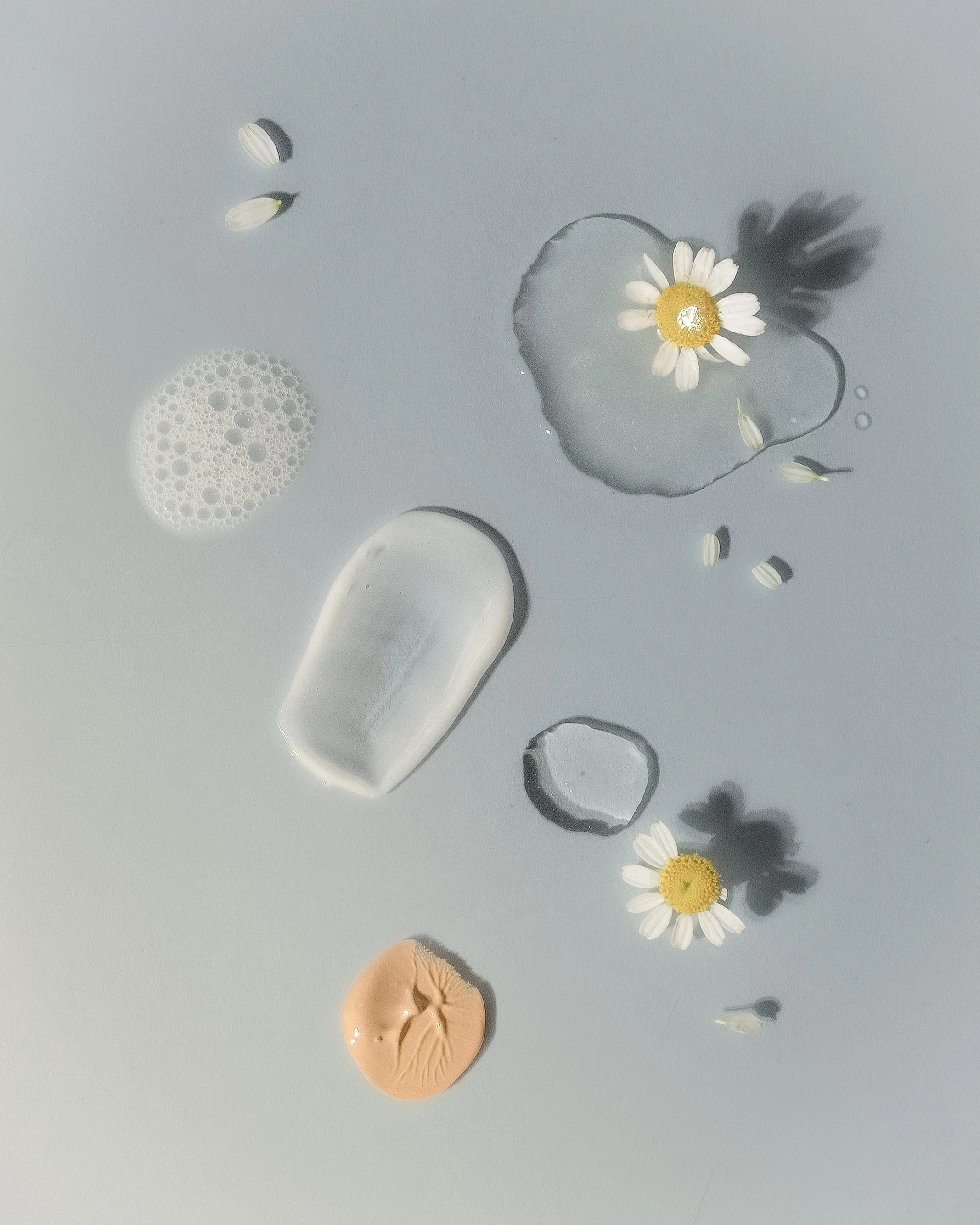 Different skincare products, including a bottle, a tube, a dollop of cream, and a soap, with daisies and water on a light gray background.