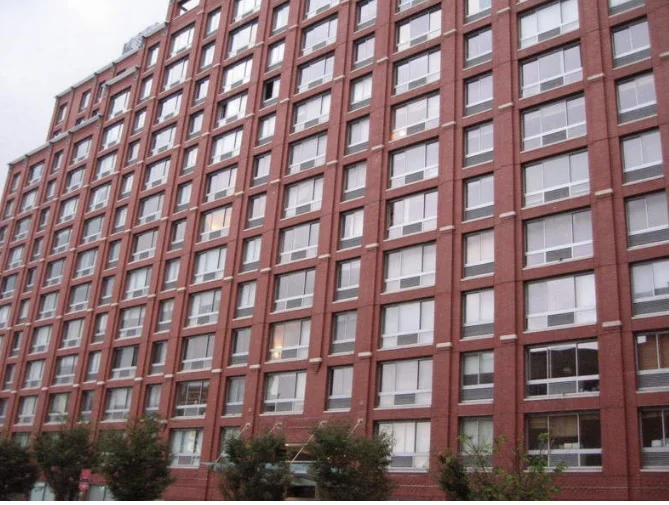 High-rise red brick apartment building with multiple windows, some with open curtains, and trees at the base.