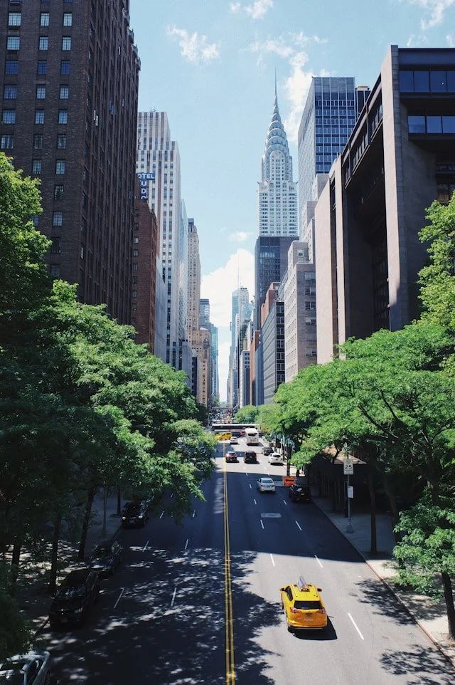 A city street in New York City with cars driving, tall buildings on both sides, green trees lining the sidewalks, and the Chrysler Building in the background under a blue sky with some clouds.