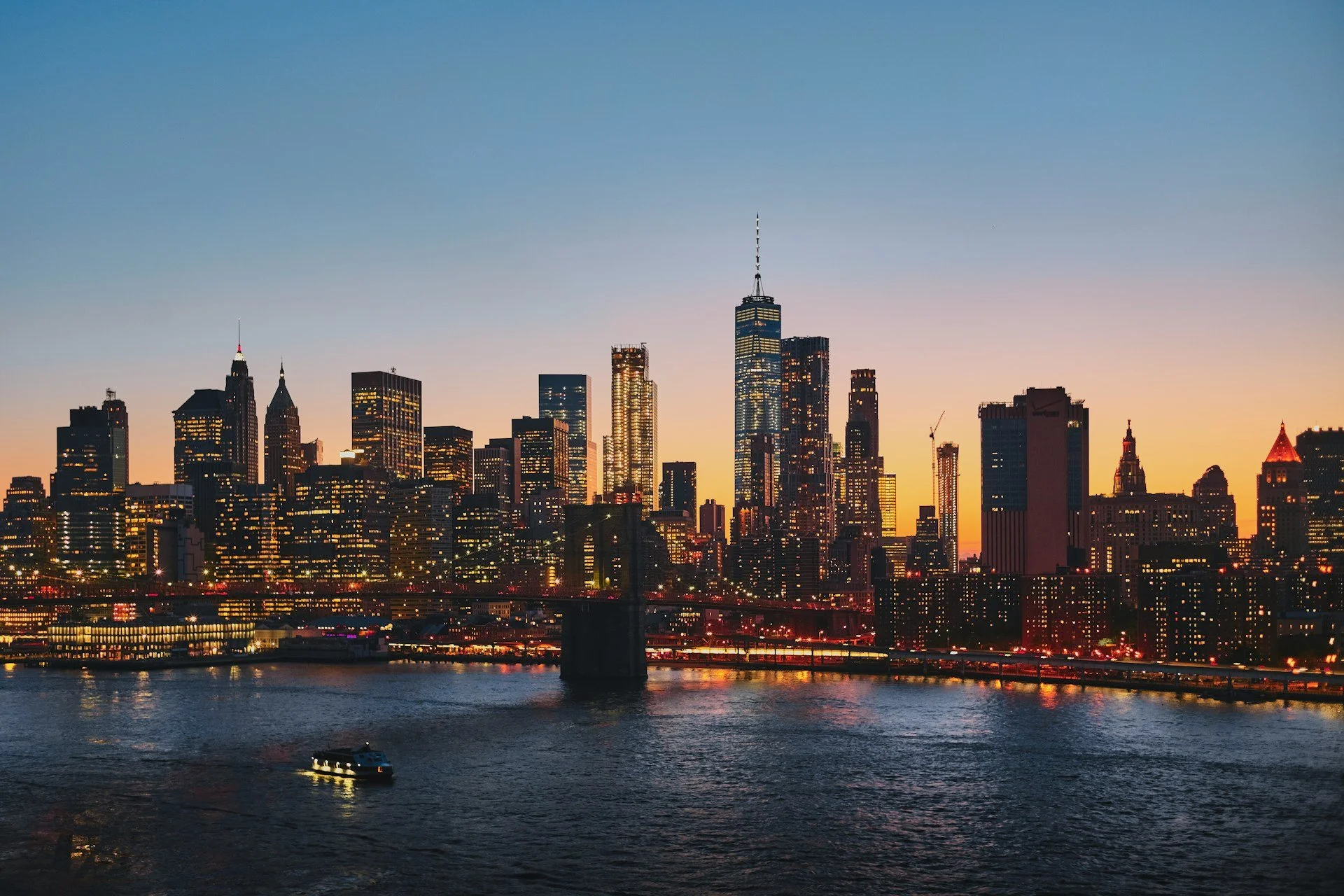 New York City skyline at sunset with illuminated skyscrapers and a boat on the river