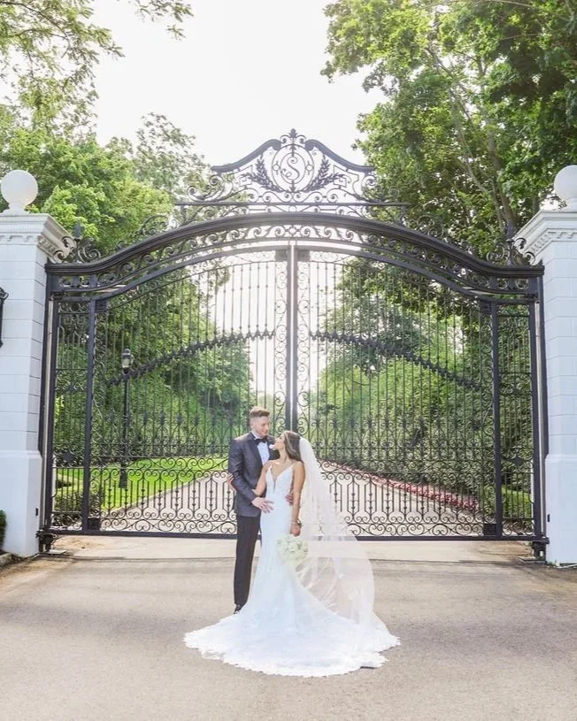 A bride and groom in wedding attire stand in front of a large, ornate black iron gate surrounded by lush green trees.