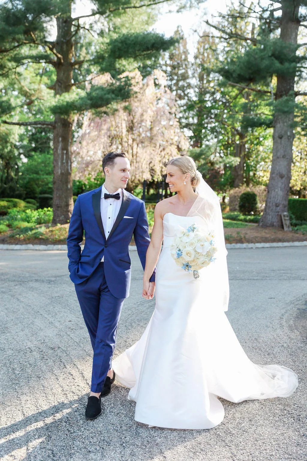 A bride and groom walking hand in hand outdoors, smiling at each other, with trees and greenery in the background. The bride wears a white wedding dress and holds a bouquet, while the groom wears a blue tuxedo and black shoes.