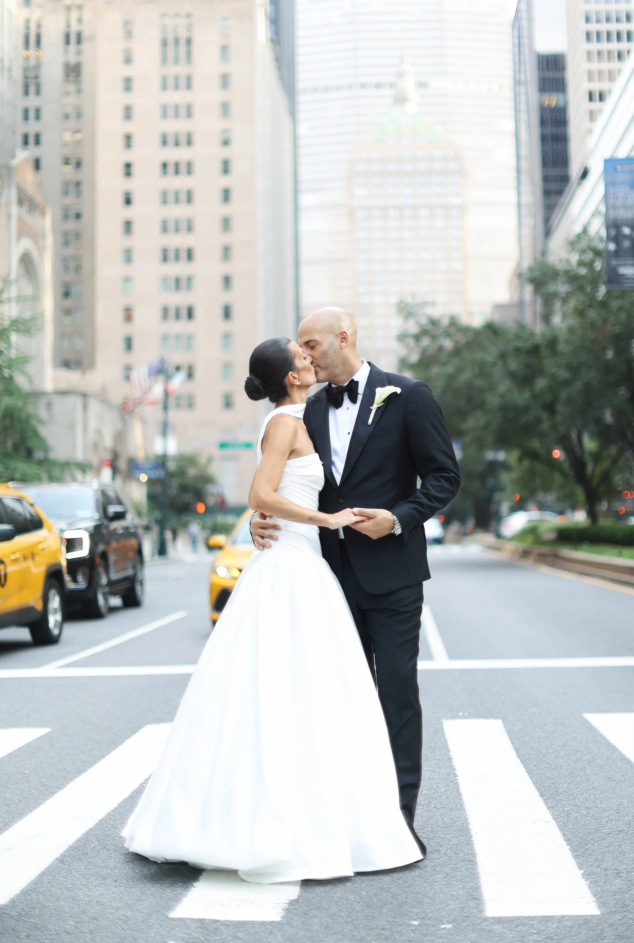 Couple in wedding attire sharing a kiss on a city street crosswalk with tall buildings and yellow taxis in the background.