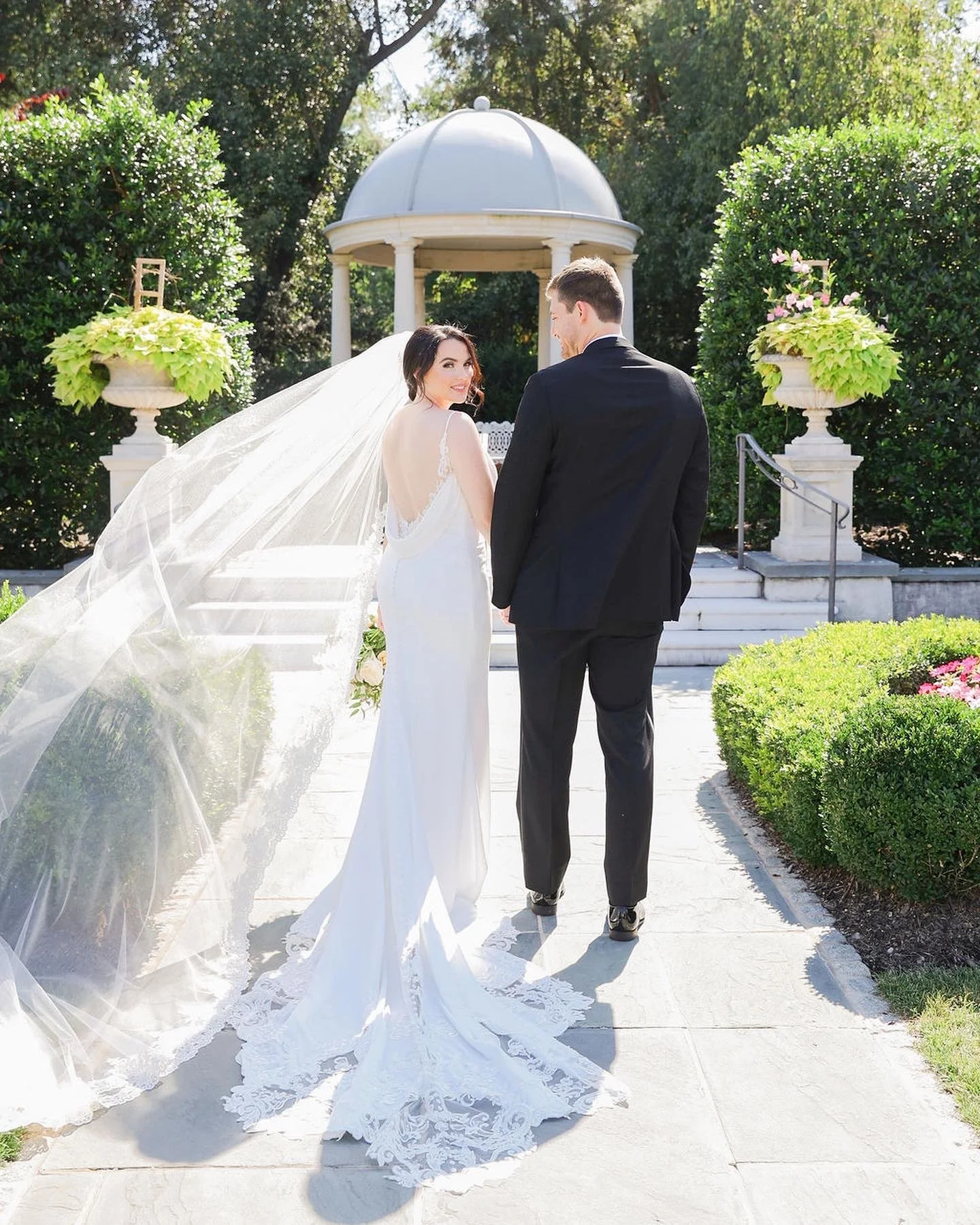 A bride and groom standing outdoors, holding hands and smiling at each other during a wedding ceremony in a garden setting with greenery and decorative planters.