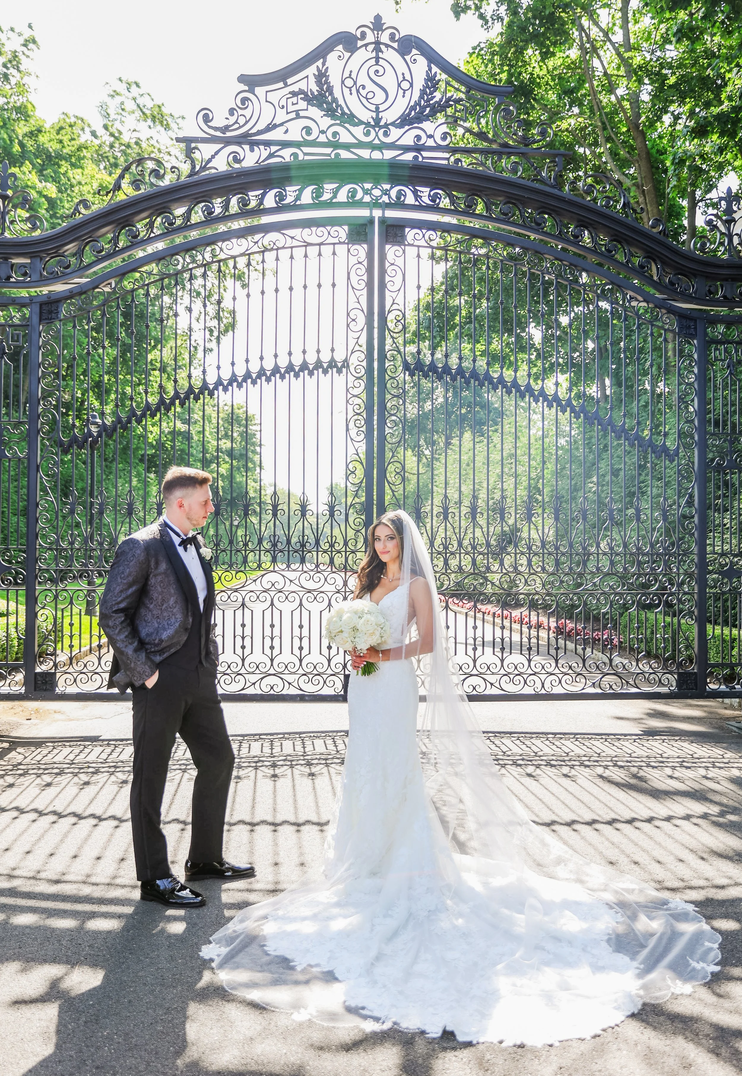 A bride in a white wedding gown and veil holding a bouquet of white flowers, standing in front of an ornate black wrought-iron gate, with a groom in a black tuxedo looking at her, outdoors on a bright sunny day with green trees in the background.