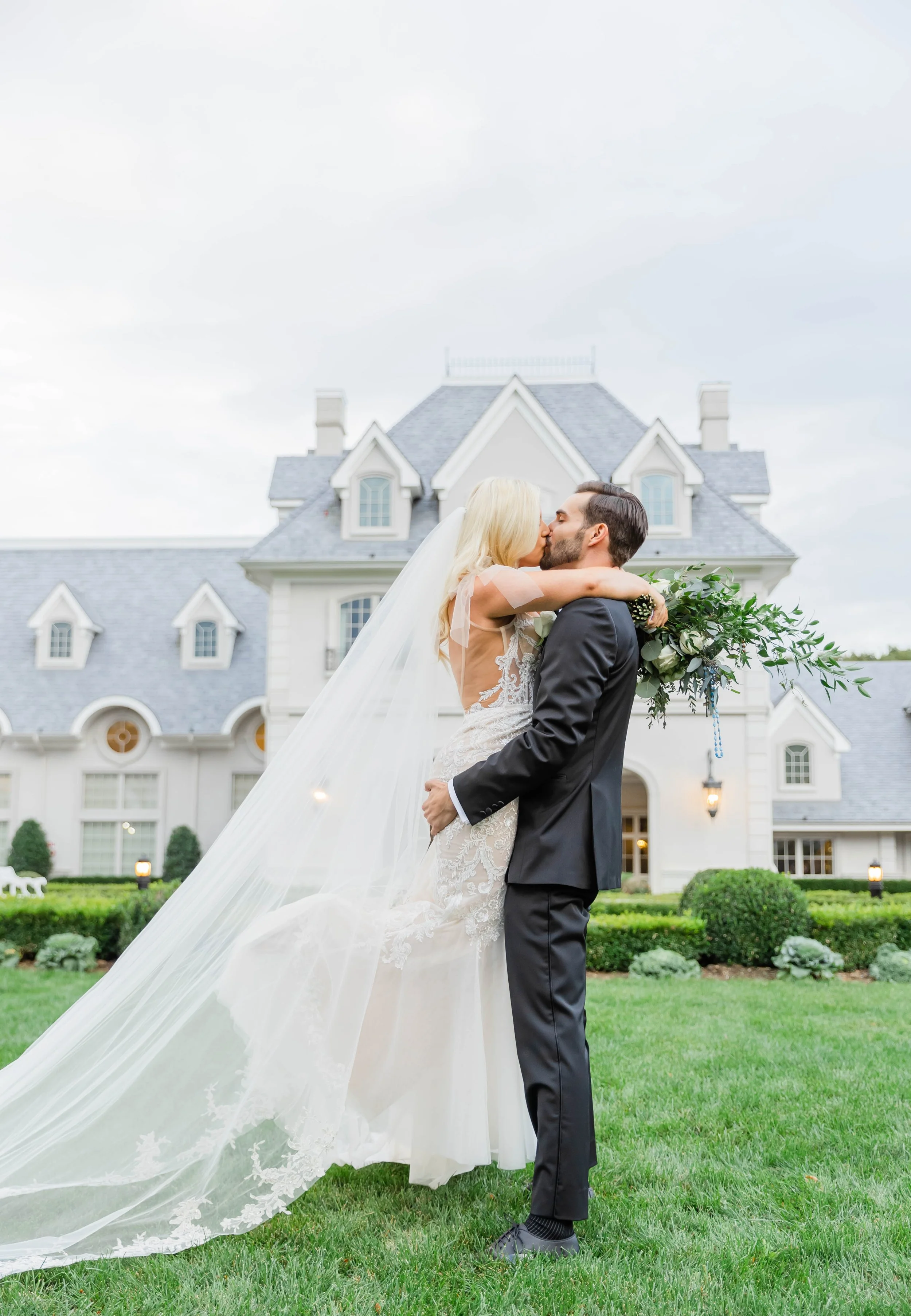A bride and groom kissing on the lawn in front of a large, white, Victorian-style house. The bride wears a white lace wedding dress and veil, and the groom wears a black suit. The bride holds a bouquet and has her arms around the groom's neck, while 