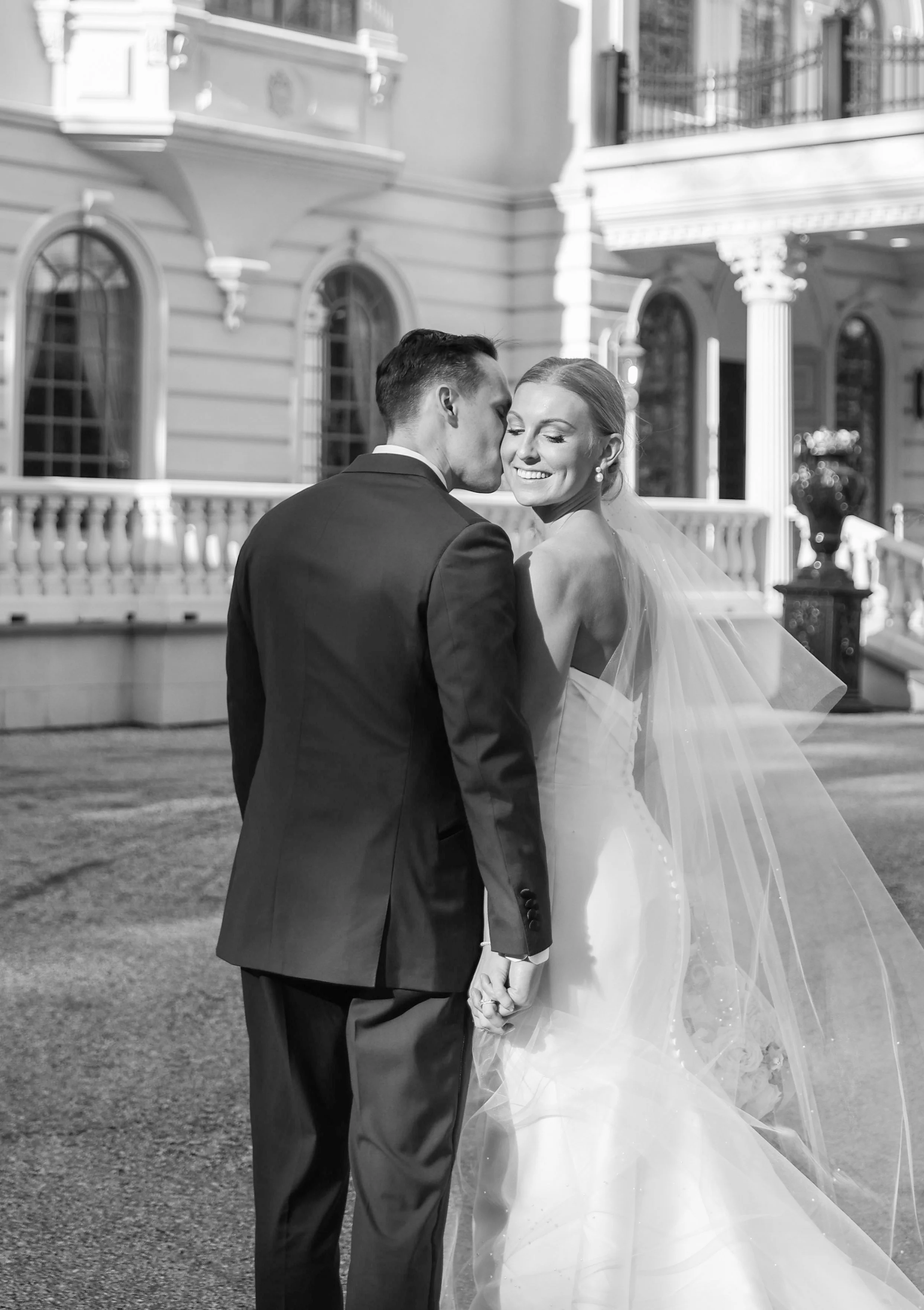 A black-and-white photo of a bride and groom holding hands outside a grand building. The groom is kissing the bride's temple, and she is smiling with her eyes closed. The bride is wearing a wedding dress with a long veil, and the groom is in a suit.