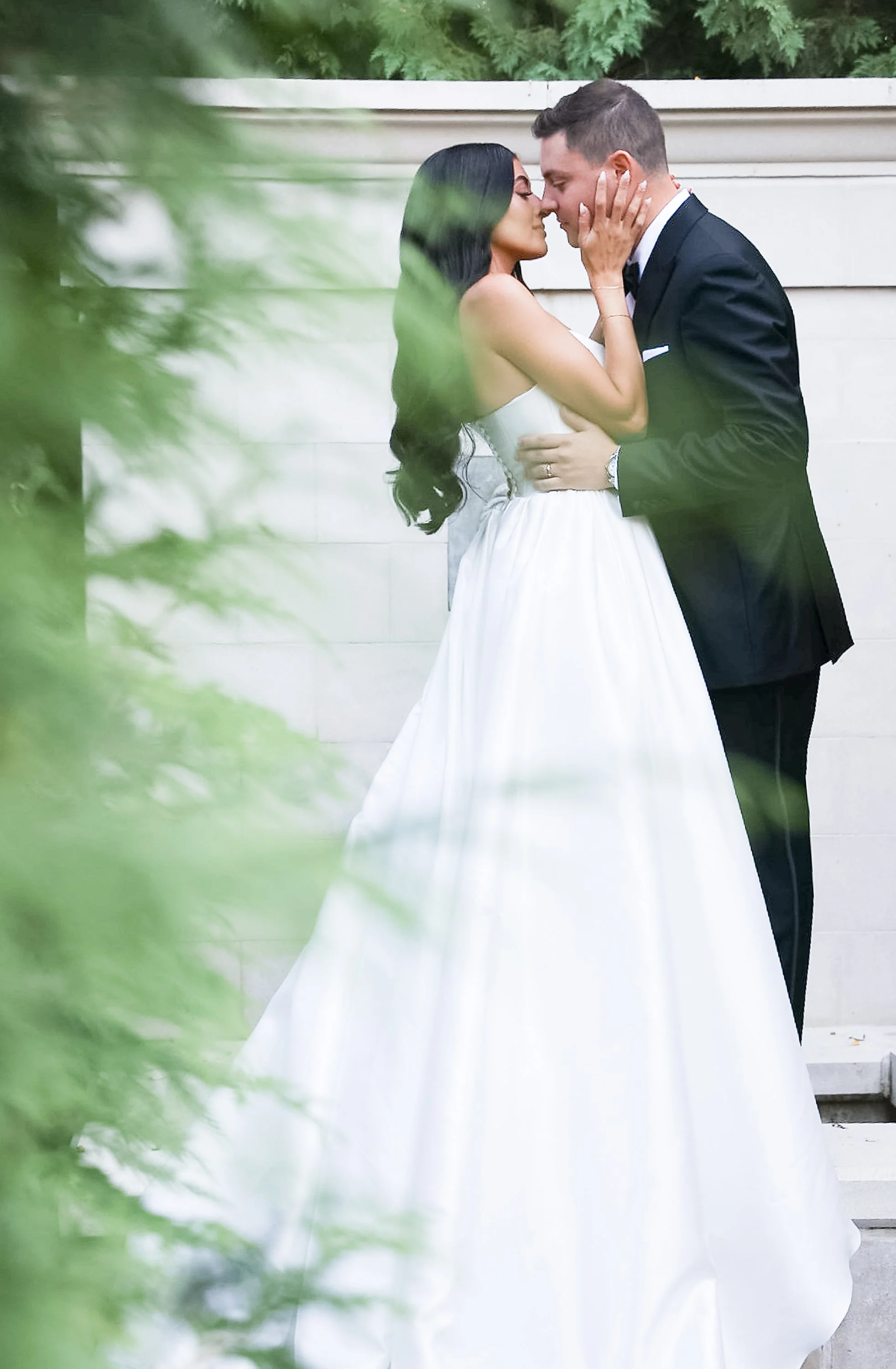A bride and groom embrace and lean in for a kiss, standing close against a neutral wall with greenery partially framing the scene.