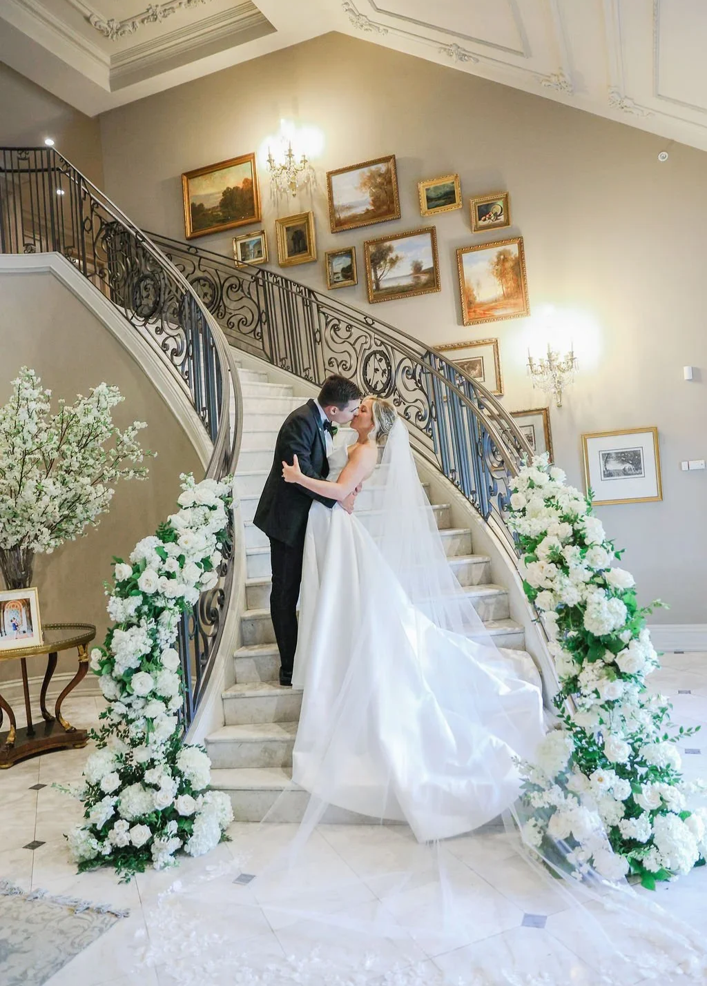 A bride and groom share a kiss on a grand staircase decorated with white floral arrangements, in an elegant interior with framed paintings on the wall and ornate chandeliers.