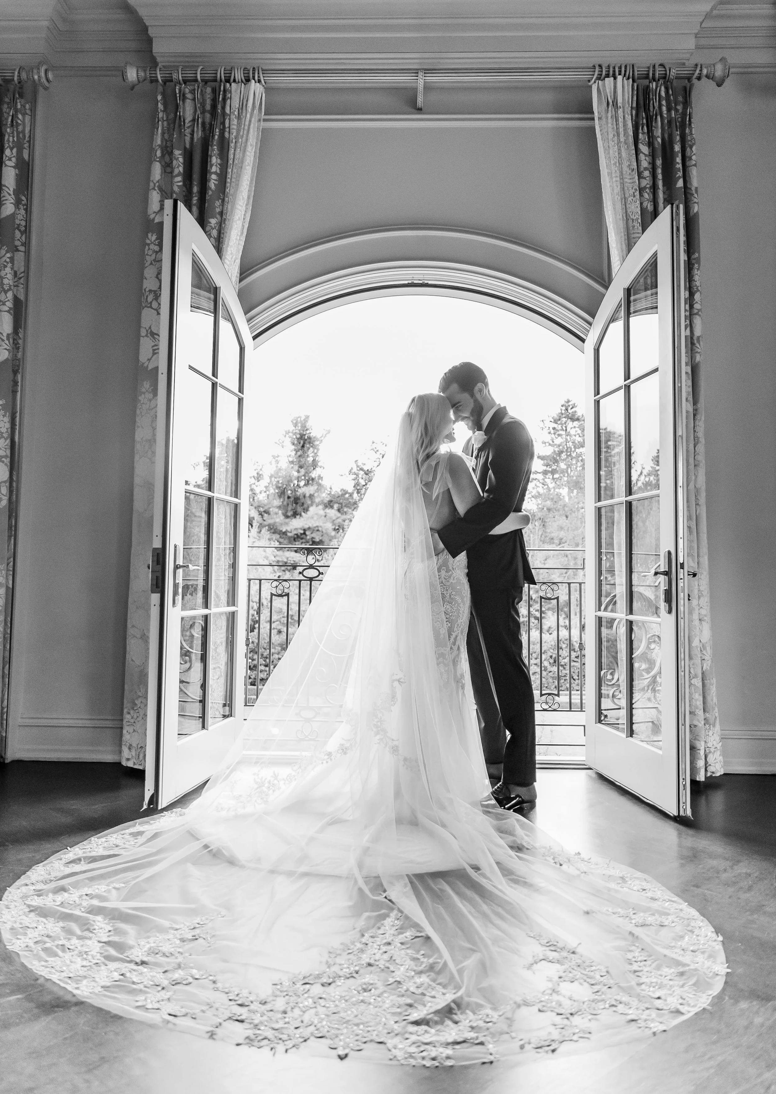 Black and white photo of a bride and groom standing close together in an open doorway on a balcony, with trees visible outside, during their wedding day. The bride wears a long, lace wedding dress with a veil, and the groom is in a dark suit with a b