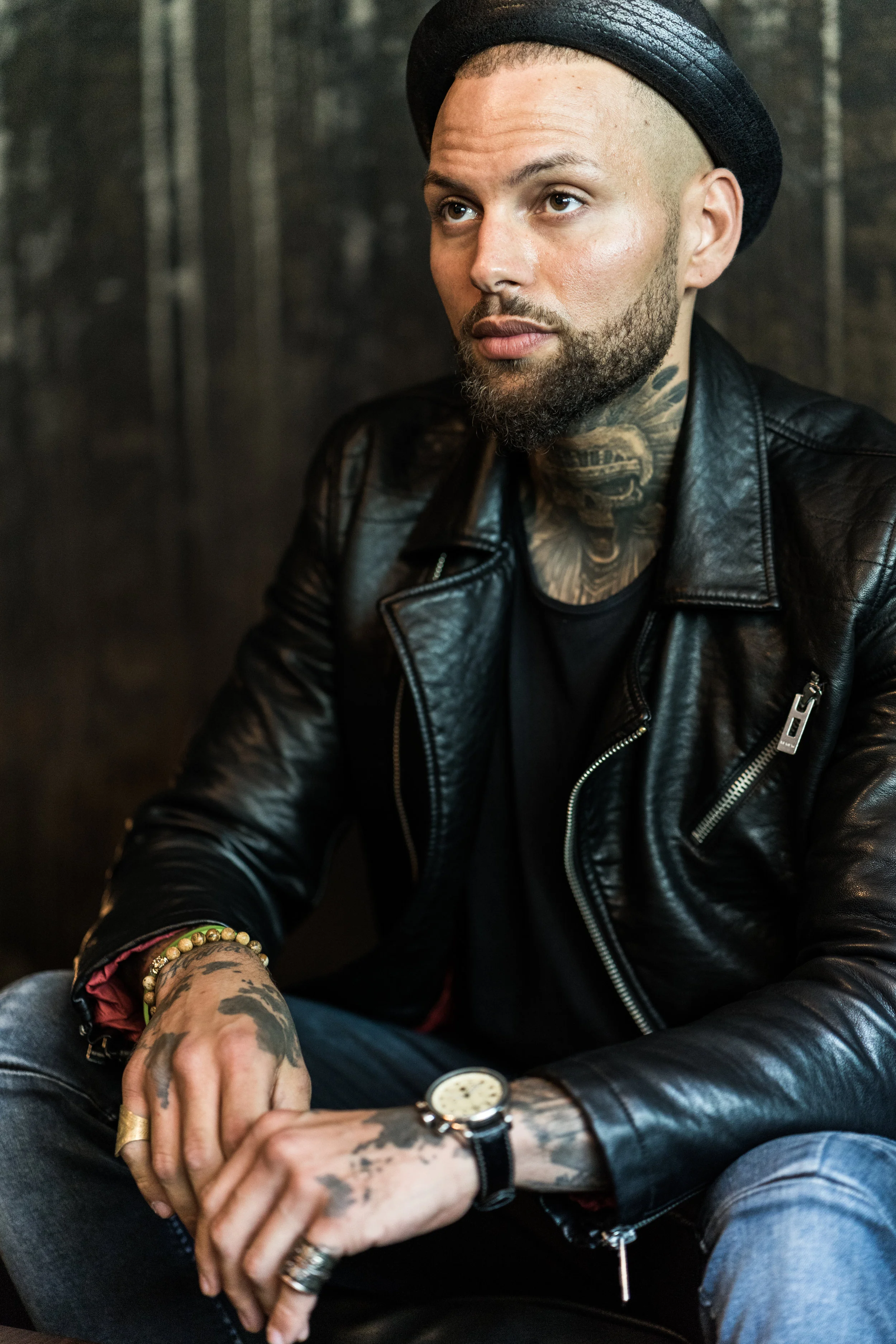Ein junger Mann mit langen Tattoos auf Händen und Hals, trägt eine schwarze Lederjacke, eine schwarze Mütze, einen Goldring, ein Armband und eine Uhr, sitzt nachdenklich vor dunklem Hintergrund.