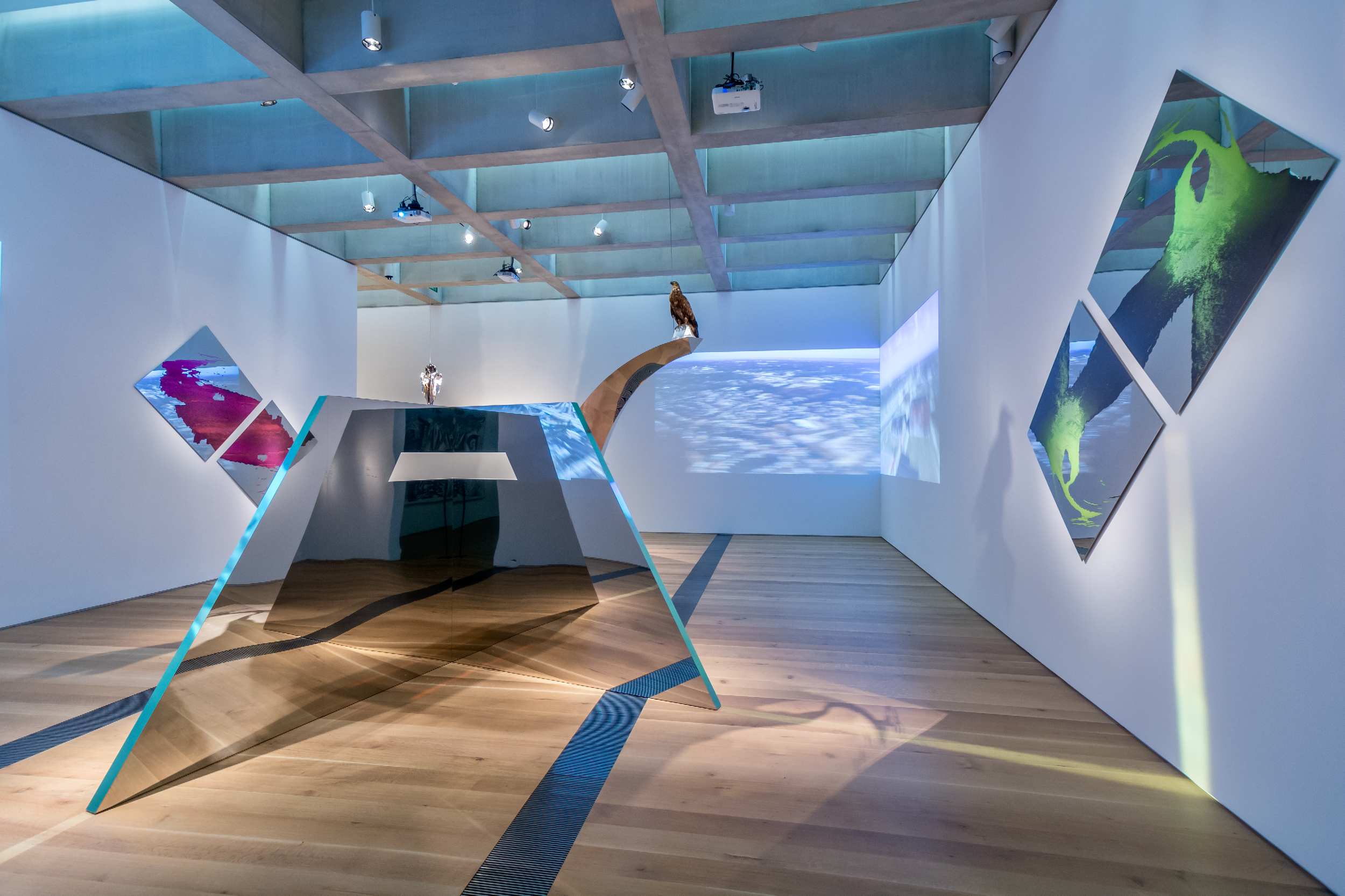 Convergence Infinité , 2016, 14’ x 40’ x 50’, mirror polished stainless steel, dichroic film, bald eagle taxidermy mount, chrome plated horse skull, acrylic box containing Mississippi and Missouri River waters, mirrored glass, archival inkjet prints on mirror polished stainless steel, and four channel HD drone footage video projection. The sculpture Apogee 1200 (foreground) is an abstracted form based on a Cahokian mound. The mirrored steel edges are coated with a dichroic film which refracts light on the edges of the sculpture. The sculpture reflects within itself, giving the illusion that one is viewing the front and the back of the work simultaneously.
