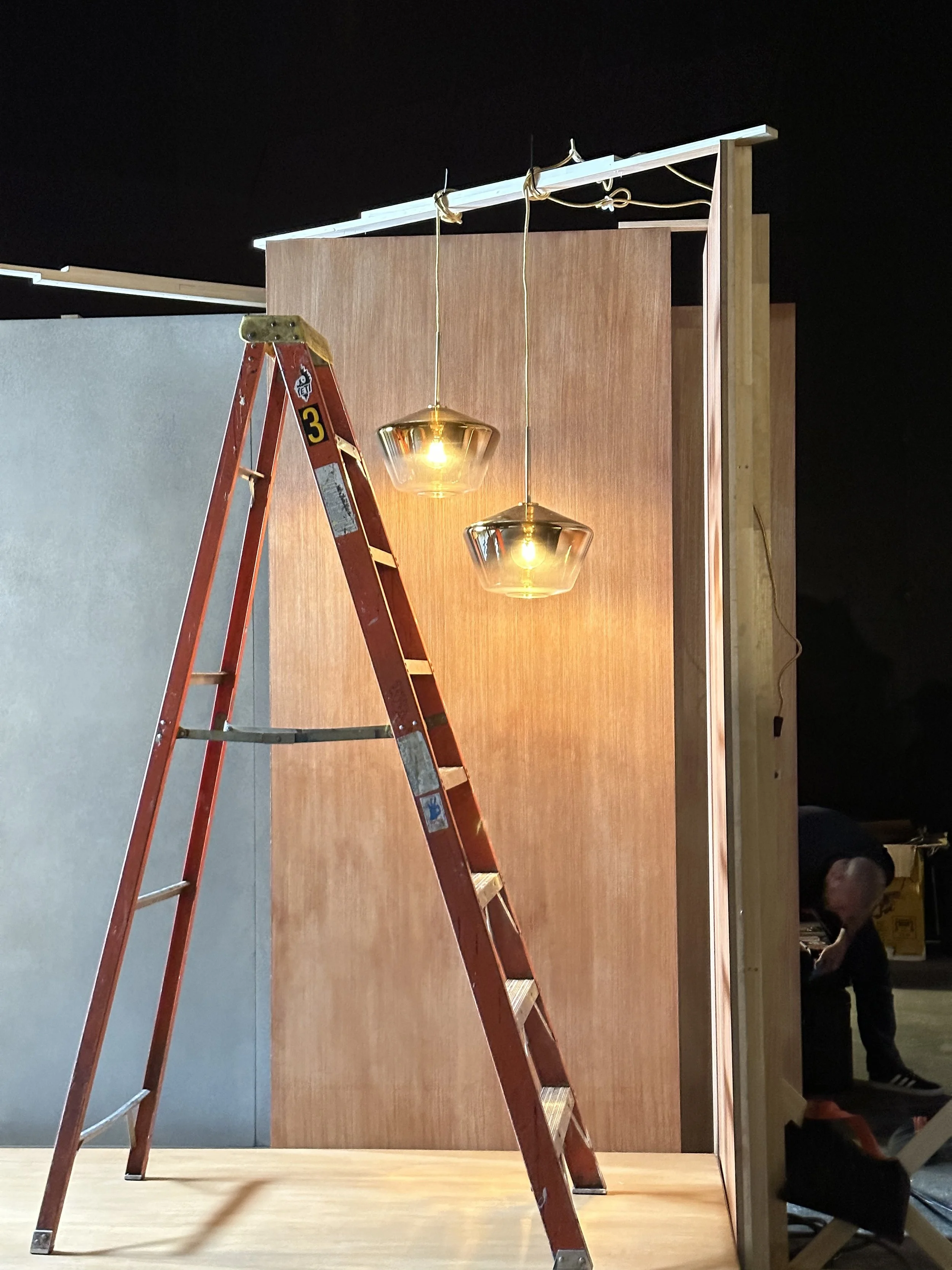 Construction scene with a red ladder, hanging light fixtures, a wooden wall, and a person working in the background.