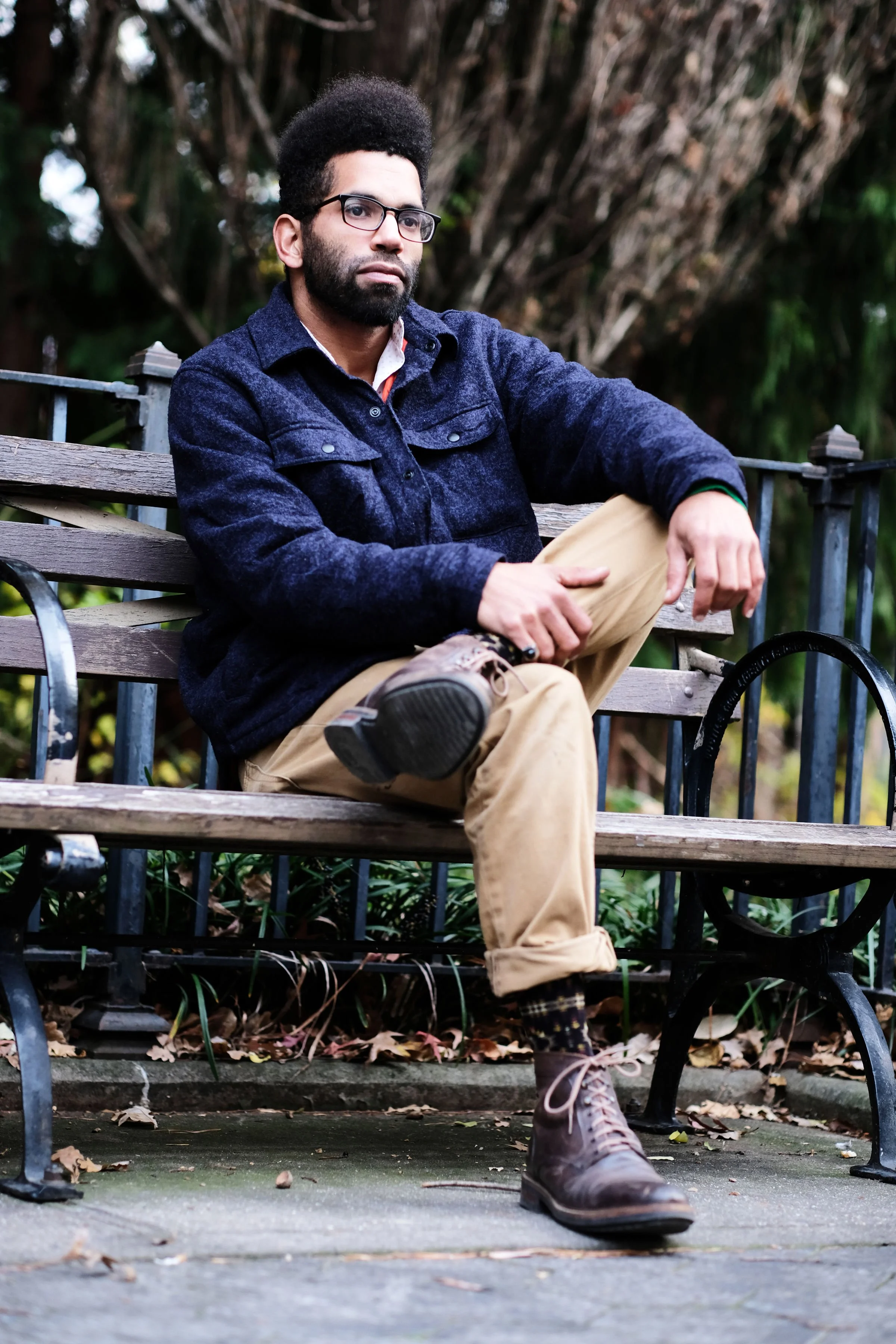 A man with glasses, a beard, and an afro hairstyle is sitting on a park bench, wearing a navy jacket, beige pants, and brown boots, with a forest background.