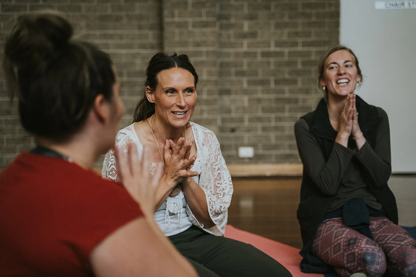 Three women sitting on yoga mats in a circle, engaged in a group discussion or meditation, in a room with brick walls.