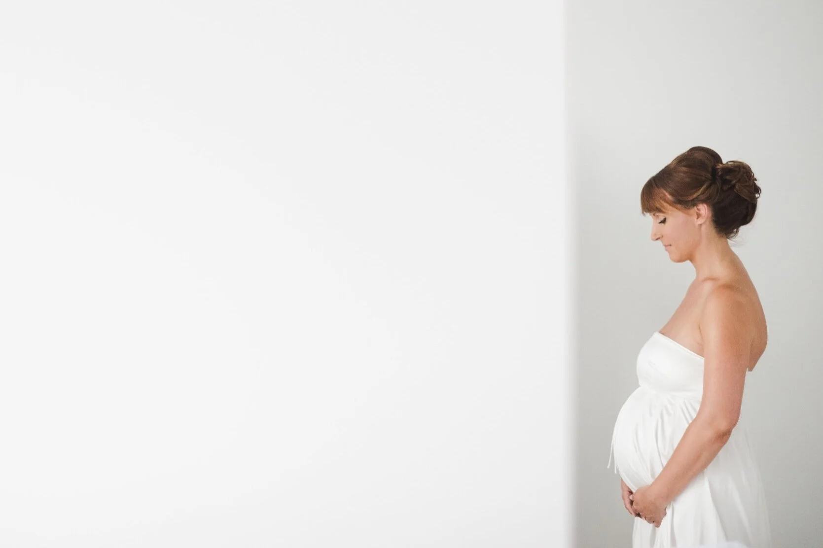 Pregnant woman in white dress standing sideways, smiling softly, with her eyes closed, against a plain white background.