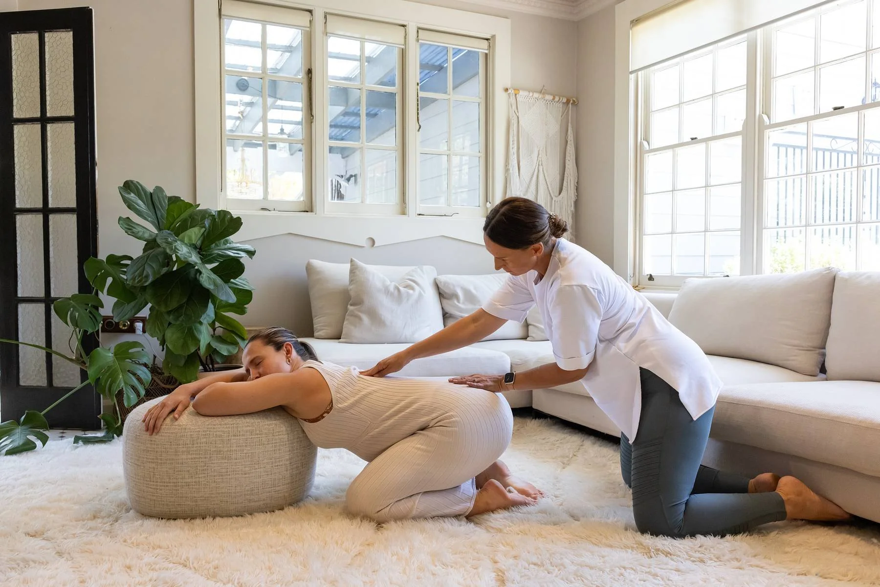 A person is receiving a massage from a therapist in a bright, cozy living room with large windows, a white sofa, and a large green plant.
