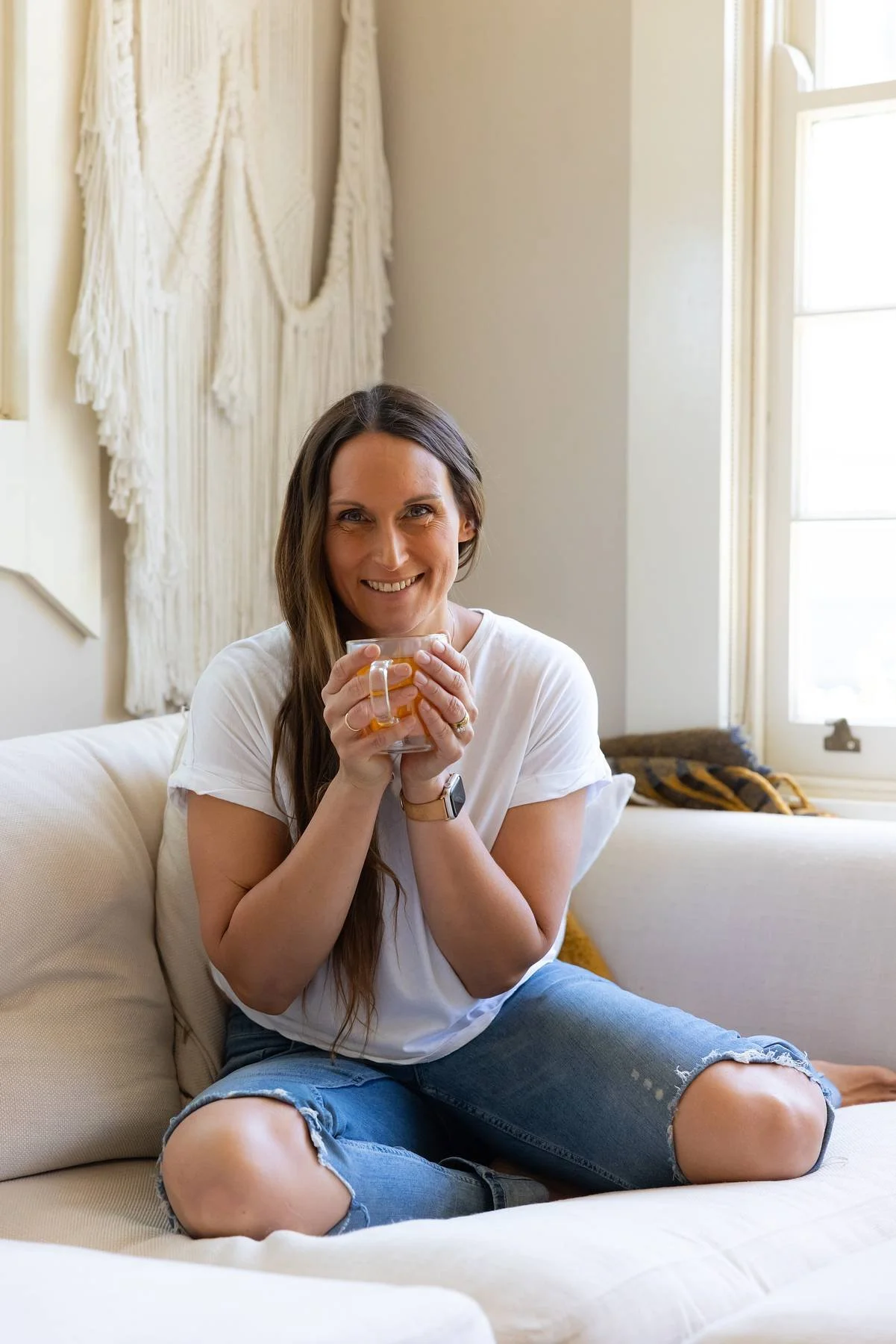 A smiling woman with long brown hair sitting cross-legged on a beige couch, holding a clear glass mug with tea, in a bright living room with sunlight streaming through a window.