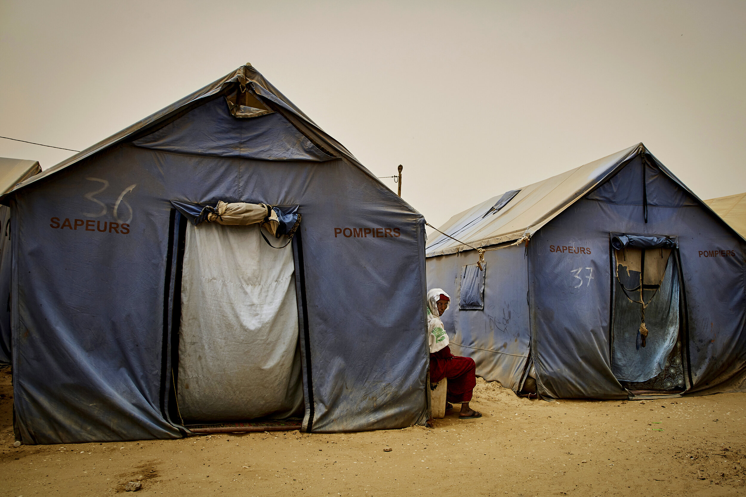   Adama Mbaye, 62, sits between two tents in Khar Yalla Camp. She along with her eldest son and his family were relocated to the camp by the government after they’d lost their home to the sea. Roughly 14 people from 3 separate families live in each t