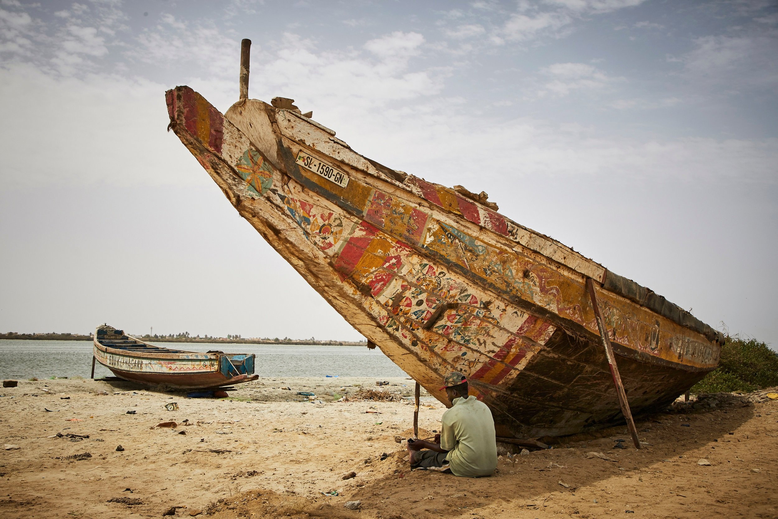  Fisherman, Daoud Diallo, sits under the bow of a boat for shade. He lives in a single room that he shares with 9 other people after his family home became inhabitable due to erosion. 