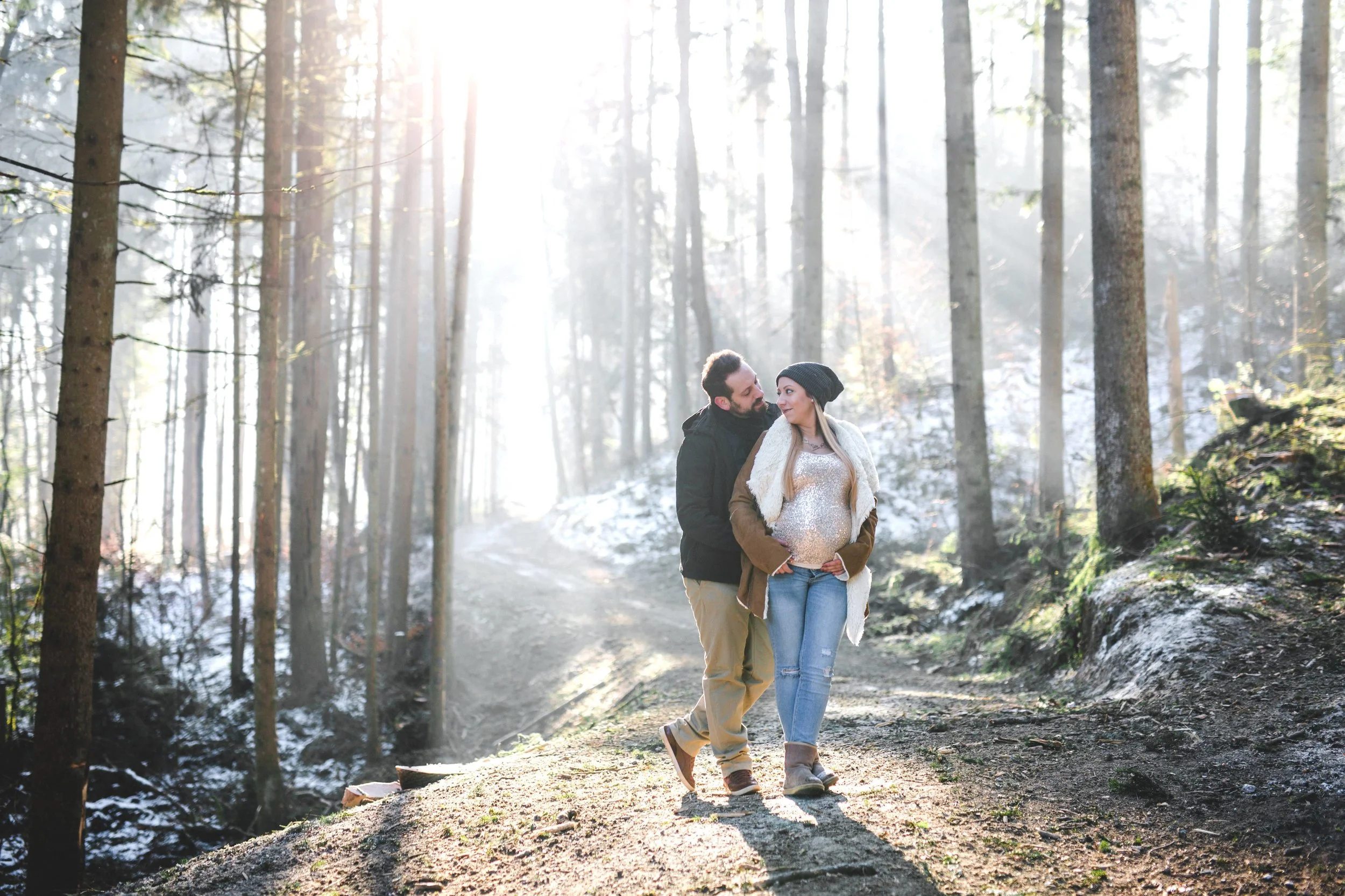 Babybauchshooting im Winter mitten im Wald bei der die werdenden Eltern auf einer Lichtung stehen und man im Hintergrund das Gegenlicht erscheint.