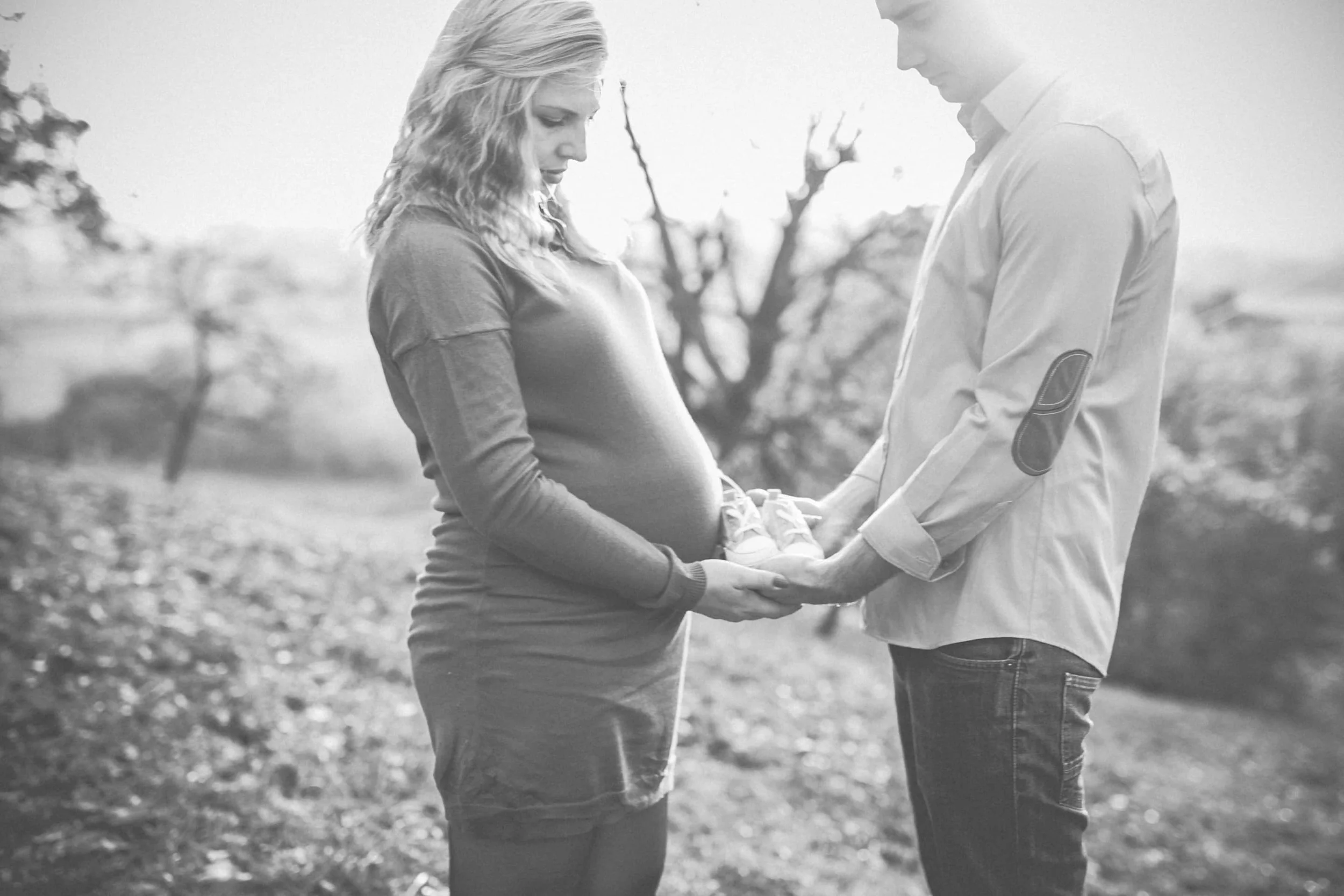 Schwarz-weiss Bild einem Schwangerschaftsshooting bei dem die werdenden Eltern sich gegenüber stehen und die Babyschuhe in der Mitte in den Händen halten.