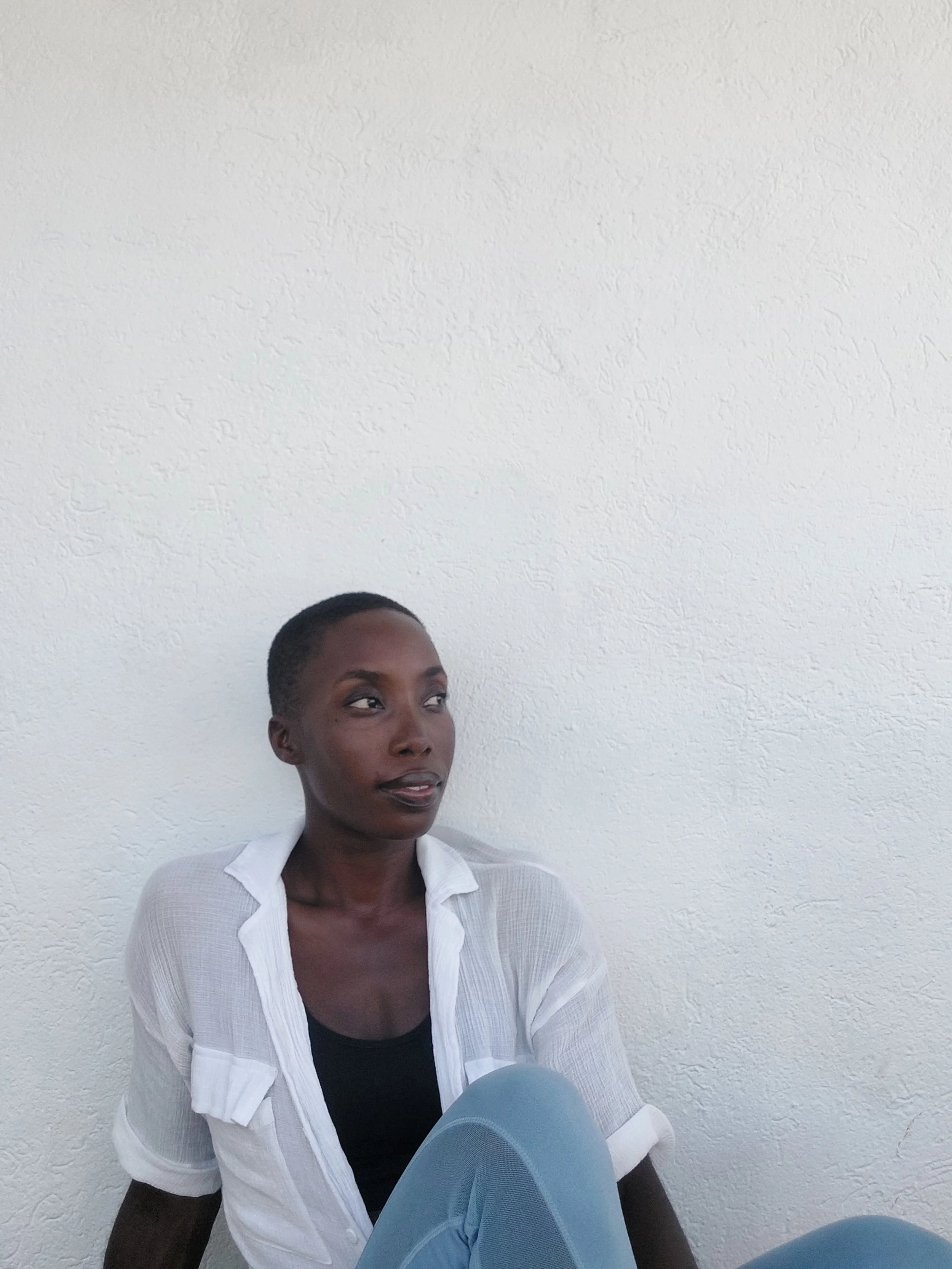 A woman with short hair sitting against a white textured wall, wearing a white button-up shirt over a black top.