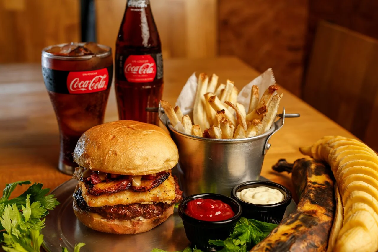 MAD Burger and Beer ¡MAD! Hay que estar bien loco para dejarlo todo y dedicarse a hacer la hamburguesa perfecta. Ilan se considera a si mismo más tico que el gallo pinto; por eso no sorprende encontrar dentro de sus creaciones maestras platos con ingredientes de los “nuestros”. Inicia su recorrido cuando viaja a Estados Unidos para certificarse como mecánico automotriz en el reconocido Universal Technical Institute, es ahí donde descubre un vacío en la gastronomía costarricense: la verdadera hamburguesa gourmet. #AVENIDACENTRAL Inspirada en un gran clásico del rock nacional de Inconsciente Colectivo. Manteniendo una identidad tica y fiel a sus sabores con el toque MAD. Pan brioche, torta de carne 100% de res fresca hecha a mano, queso artesanal empanizado, plátano maduro, chicharrón bites y nuestra salsa secreta ahumada.