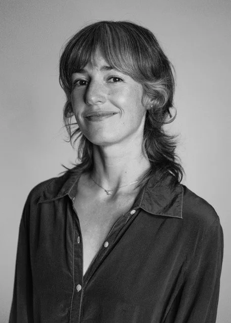 In this black and white photo, Tara looks toward the camera with a gentle smile. Her button-up blouse is unbuttoned at the top to create a V neck. Her hair is down, framing her face.