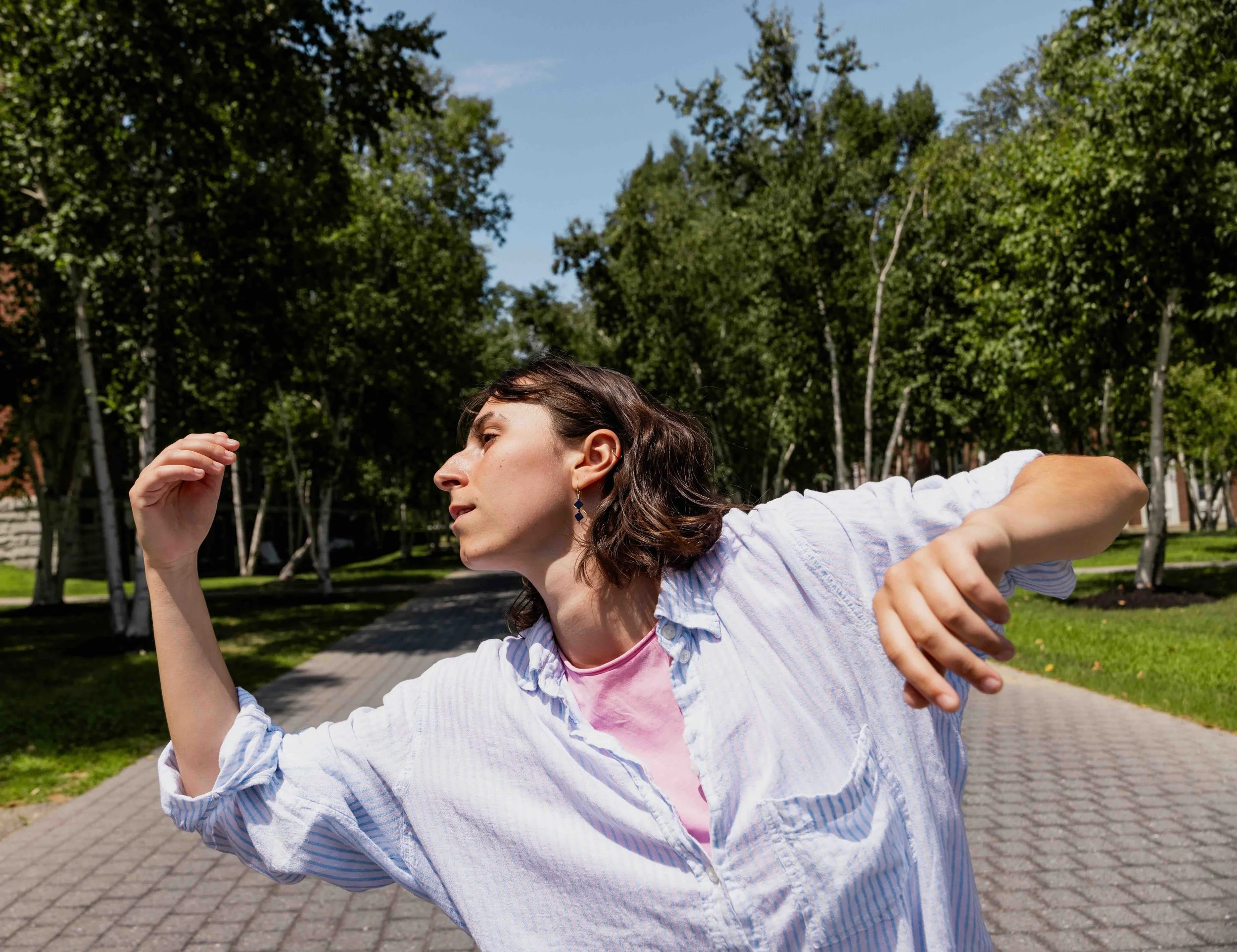 A picture of Maeve dancing. She is outside on a sunny day wearing a loose, off-white buttonup. Her brown hair is down, just brushing her shoulder; her face is turned toward her right.