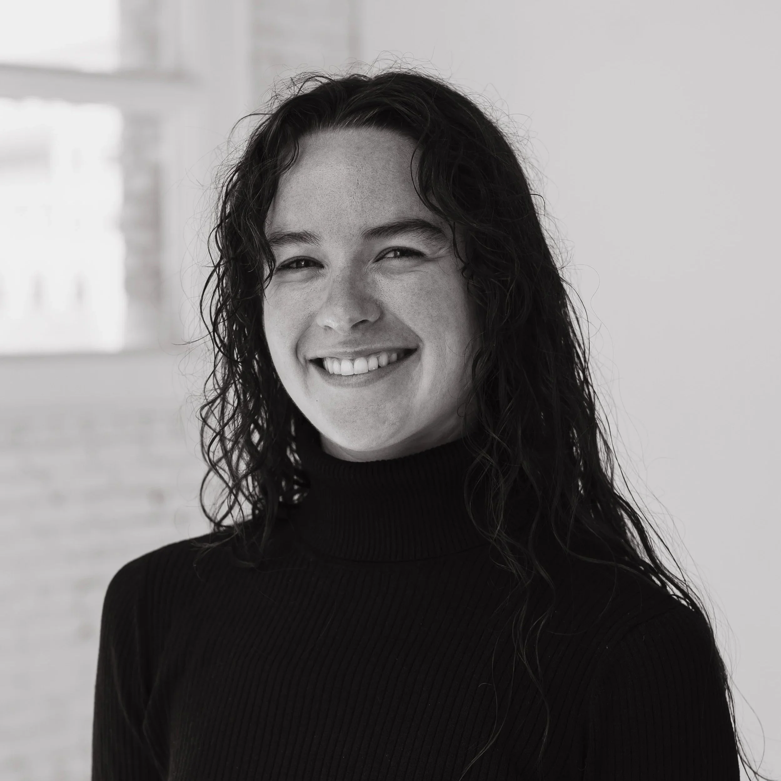 In this portrait, Kara looks to the camera wearing a black turtle neck. Her dark curly hair is down. The picture is black and white.