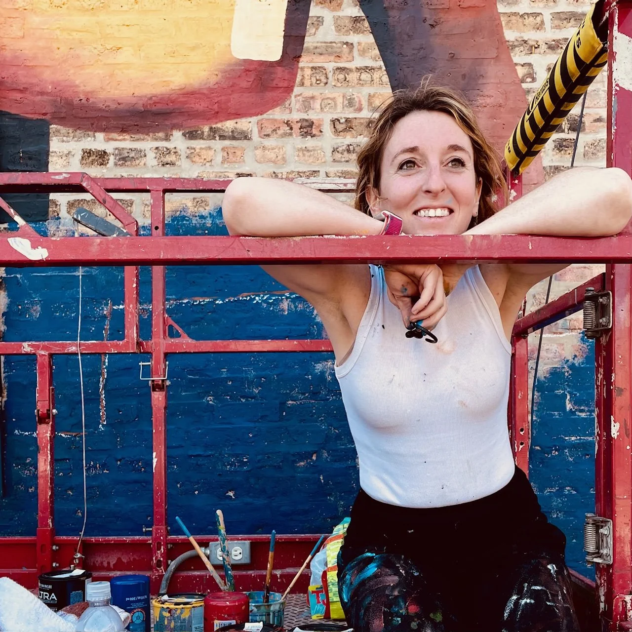 Ryan Tova Katz smiling while kneeling on a lift, working on a brick wall mural in Chicago
