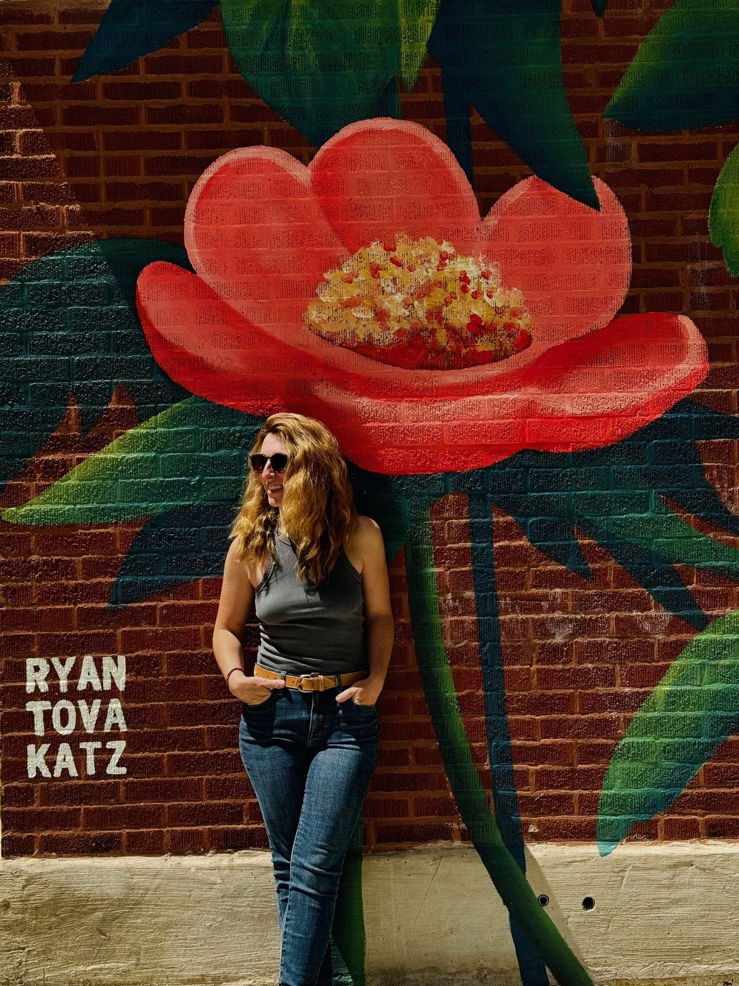 Chicago Mural Artist, Ryan Tova Katz, standing in front of one of her flower paintings on a brick wall