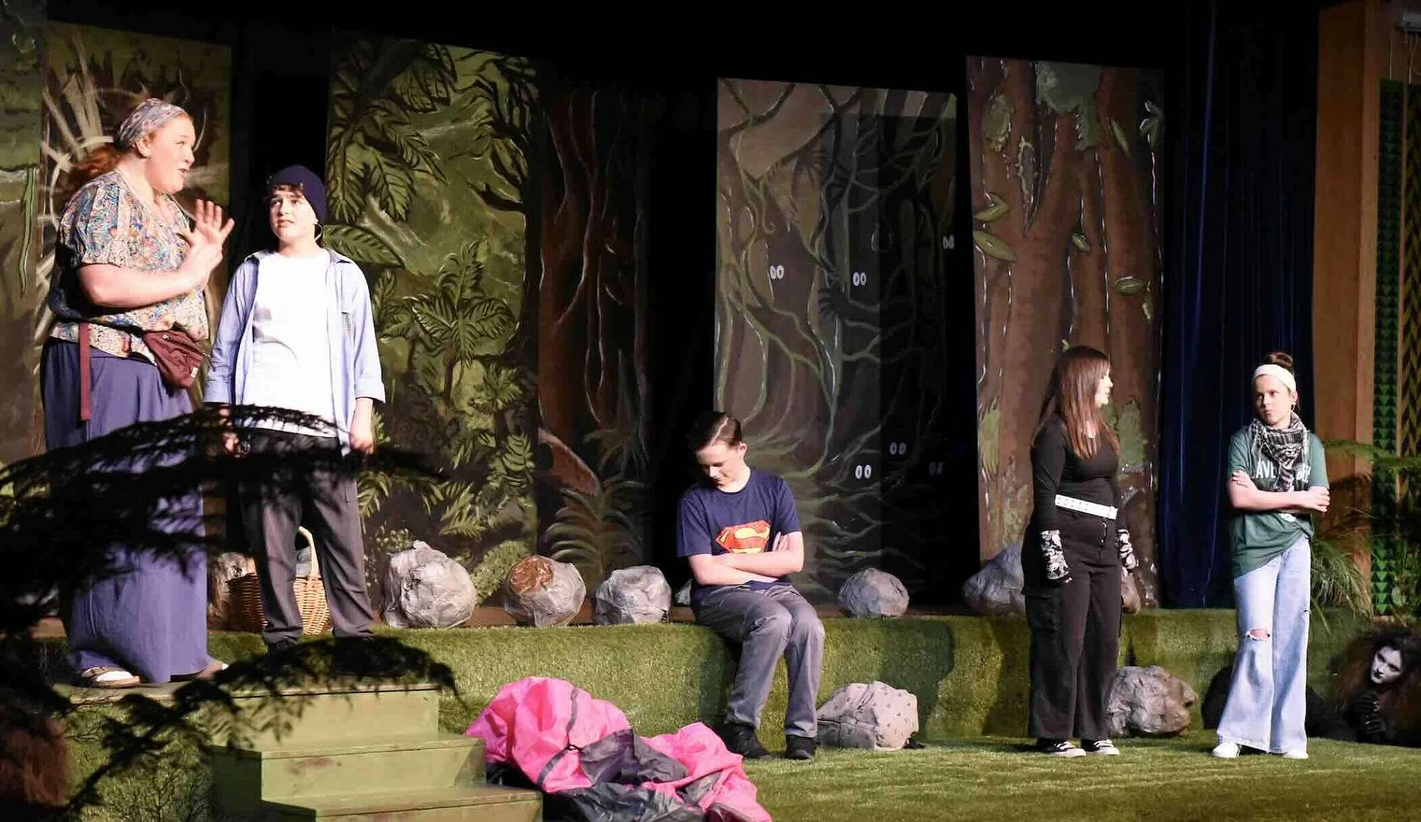 A stage with six performers on it. The stage is decorated to look like a forest with painted flats, fake grass and rocks in tiers.