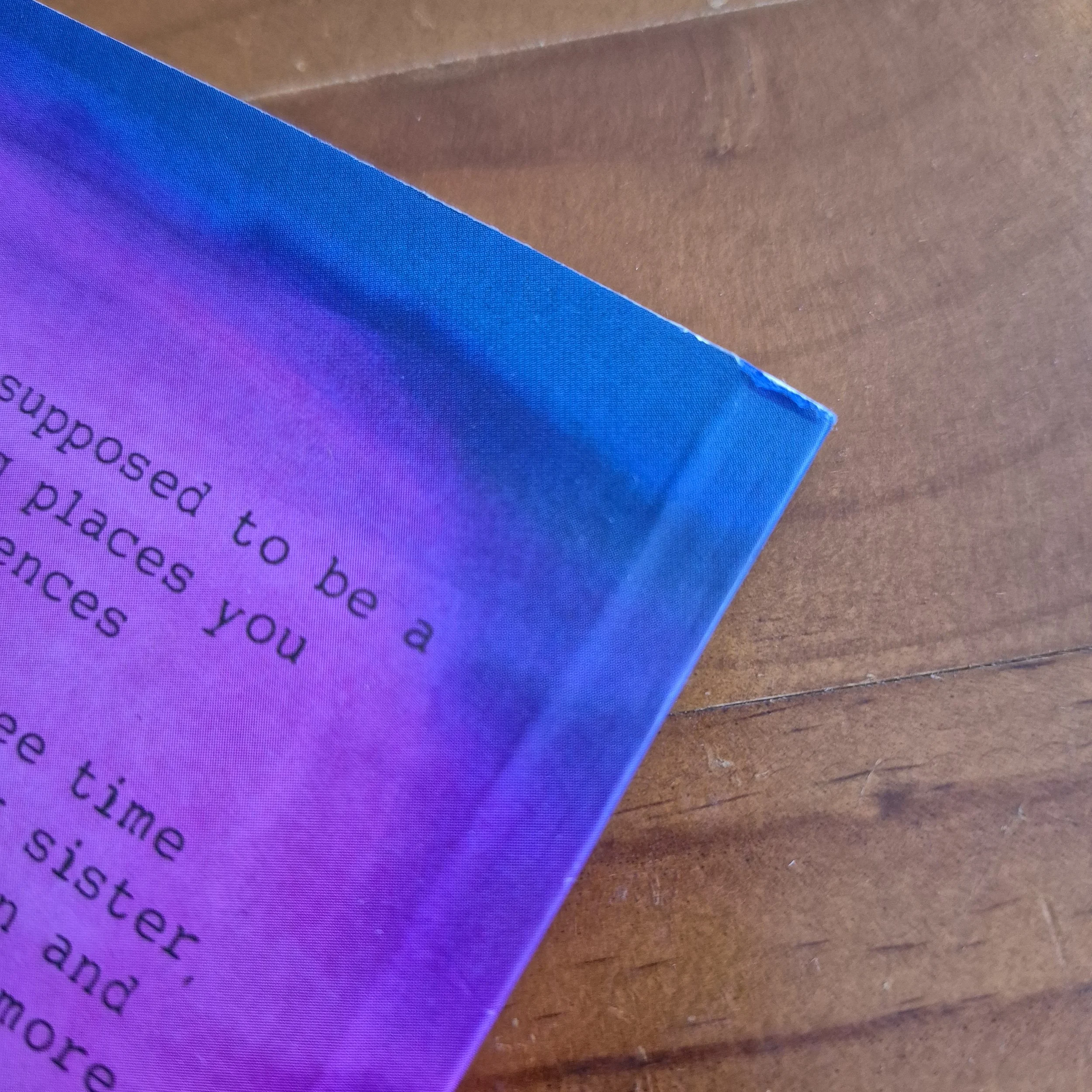 The corner of a book against a wooden background. The cover has a tiny tear near the spine on the back.
